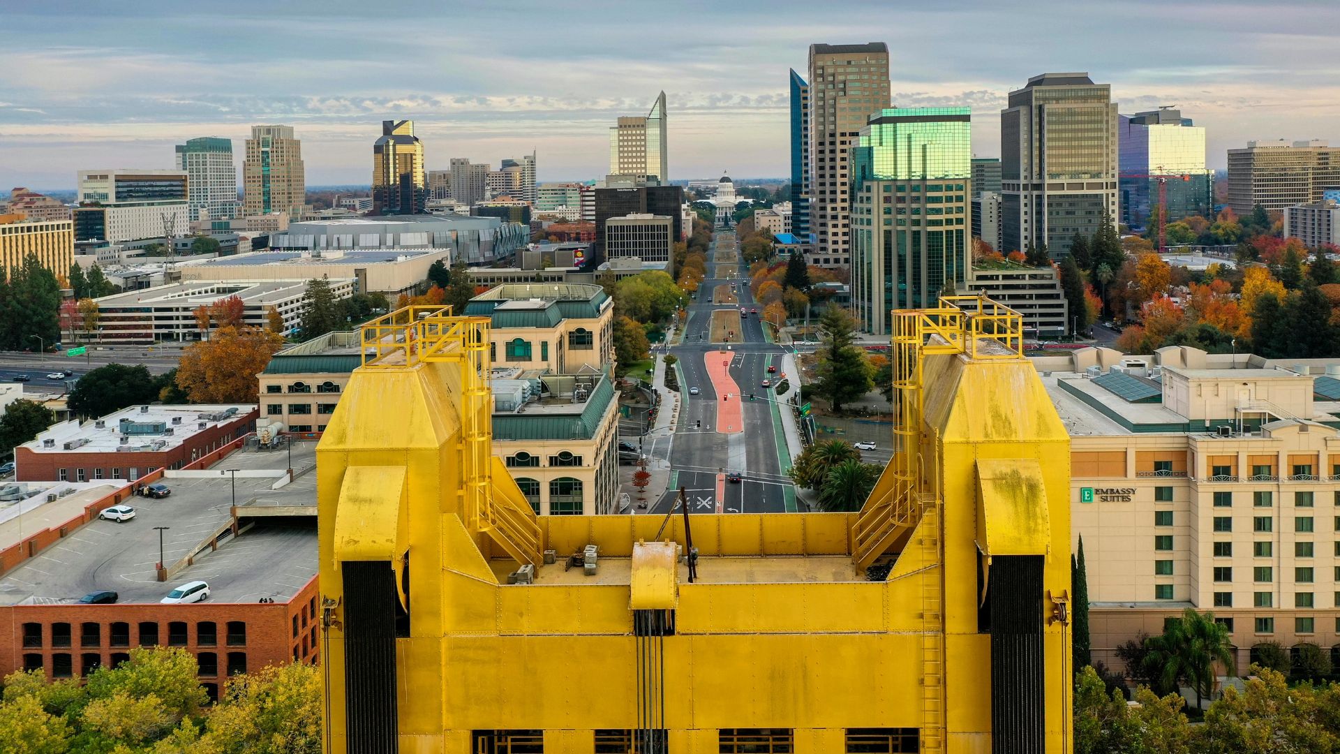 An aerial view of downtown Sacramento, California, featuring the iconic yellow Tower Bridge in the foreground, leading to a street extending into the cityscape with various buildings and the city skyline under an overcast sky. 