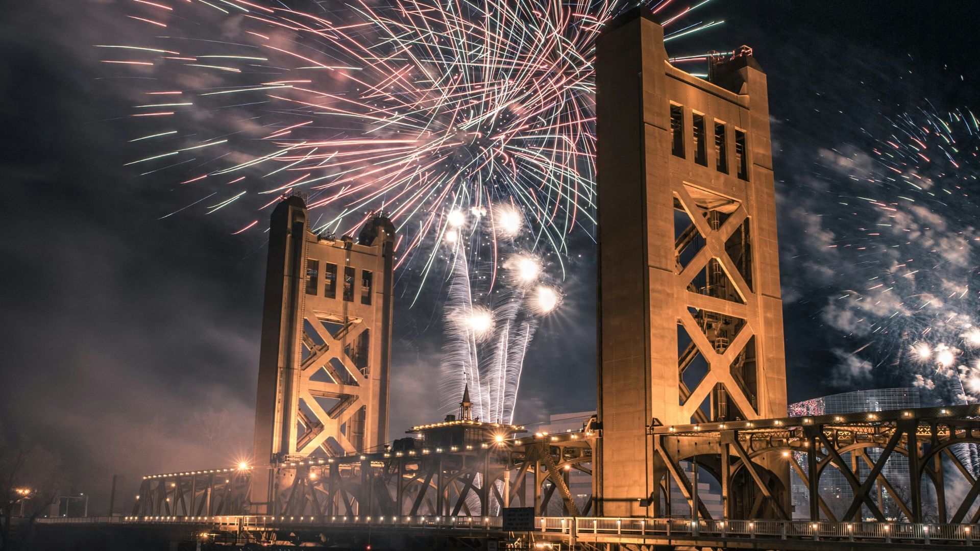 A nighttime long-exposure photograph captures the iconic Tower Bridge in Sacramento, California, illuminated in golden light, with vibrant fireworks exploding in the dark sky above and around its two distinctive towers. The Sacramento River flows beneath the bridge, reflecting the lights and fireworks, while spectators can be seen gathered on the riverbanks and docks below.