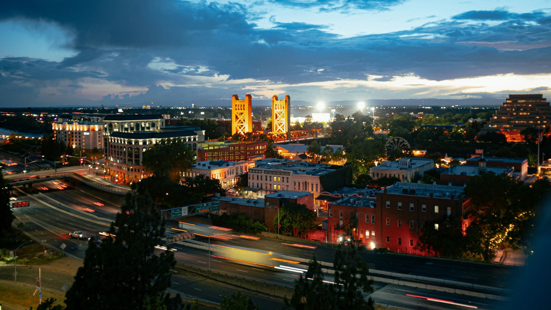 An elevated, wide-angle shot of the Sacramento, California skyline at dusk or night, showcasing the illuminated gold Tower Bridge prominently in the center, with city buildings, a Ferris wheel, and streaks of light from vehicle traffic on a highway in the foreground under a dramatic cloudy sky.