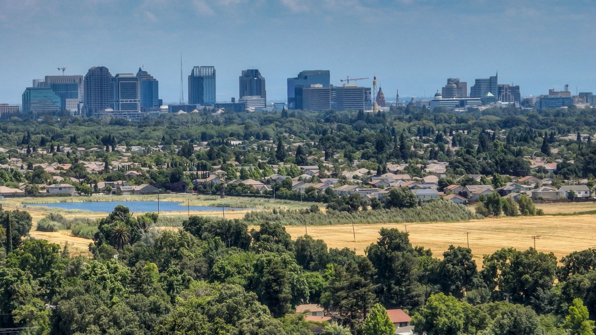 A panoramic view of Sacramento, California, showcasing a skyline of modern buildings in the distance, surrounded by a sprawling residential area with numerous houses and abundant green trees, under a partly cloudy blue sky. A body of water and a field of golden-brown vegetation are visible in the foreground.