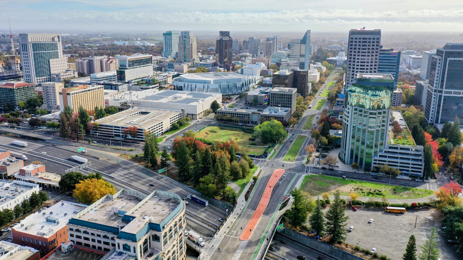 An aerial view of the urban landscape of Sacramento, California, featuring a mix of modern high-rise buildings, green spaces, and a complex network of roads and highways with vehicles. A prominent bridge with a distinct orange lane crosses over a highway, and the city skyline is visible under a partially cloudy sky.