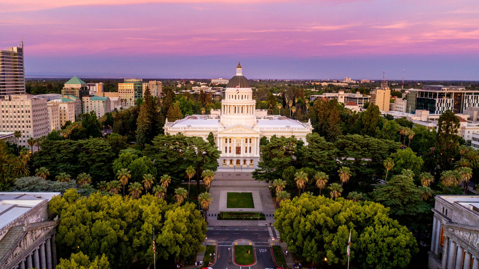 An aerial view at sunset of the California State Capitol building in Sacramento, a grand white structure with a prominent dome, surrounded by lush green trees and other city buildings under a vibrant pink and purple sky.