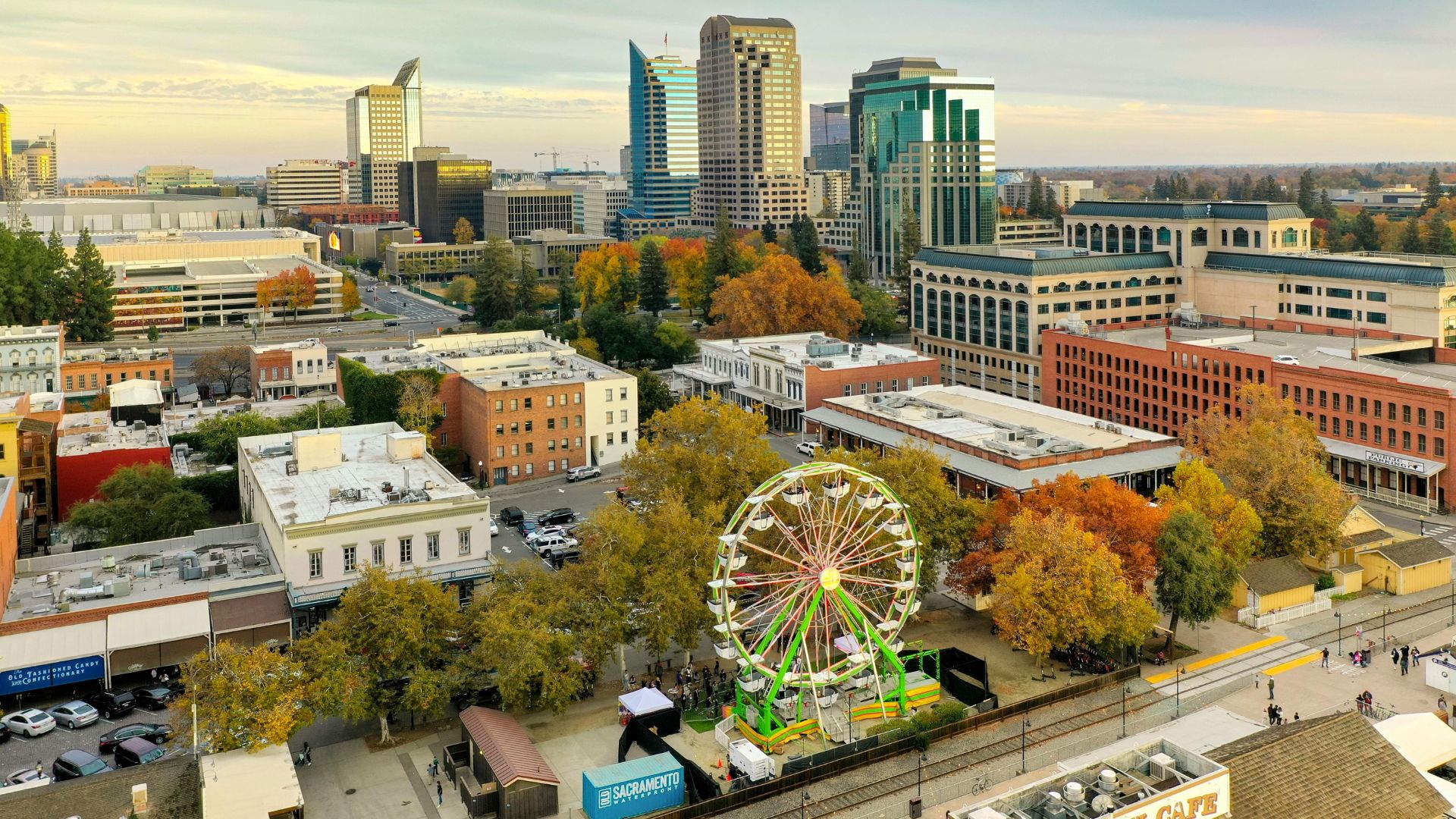 An aerial view of the Sacramento, California skyline at sunset, featuring a prominent Ferris wheel in the foreground surrounded by autumn-colored trees and various buildings, with modern skyscrapers in the background.