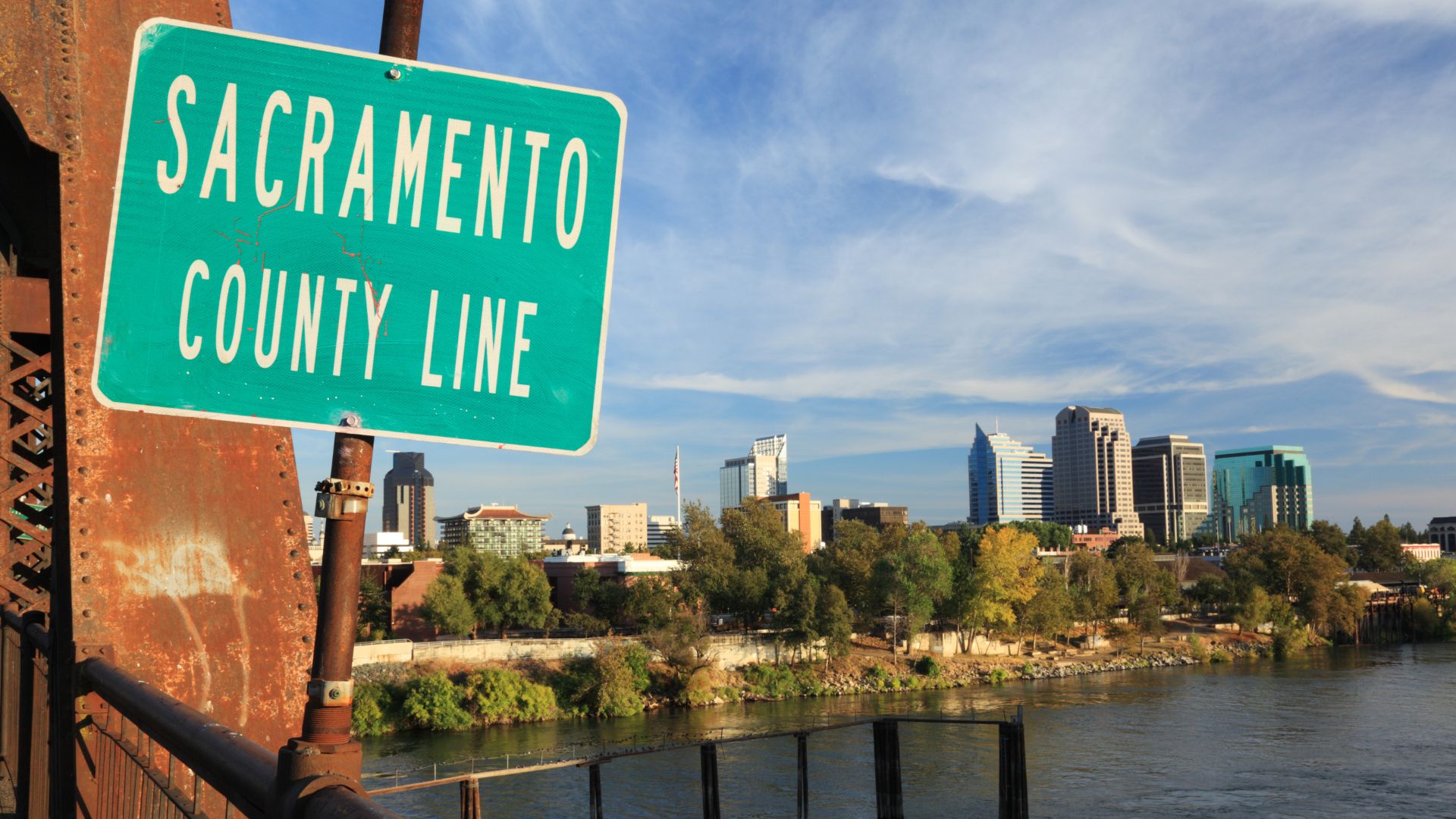 A green rectangular sign on a rusty metal post reads "SACRAMENTO COUNTY LINE" in white capital letters, set against a backdrop of a city skyline with modern buildings and a river in the foreground under a partly cloudy blue sky, indicating the boundary of Sacramento County, California. 