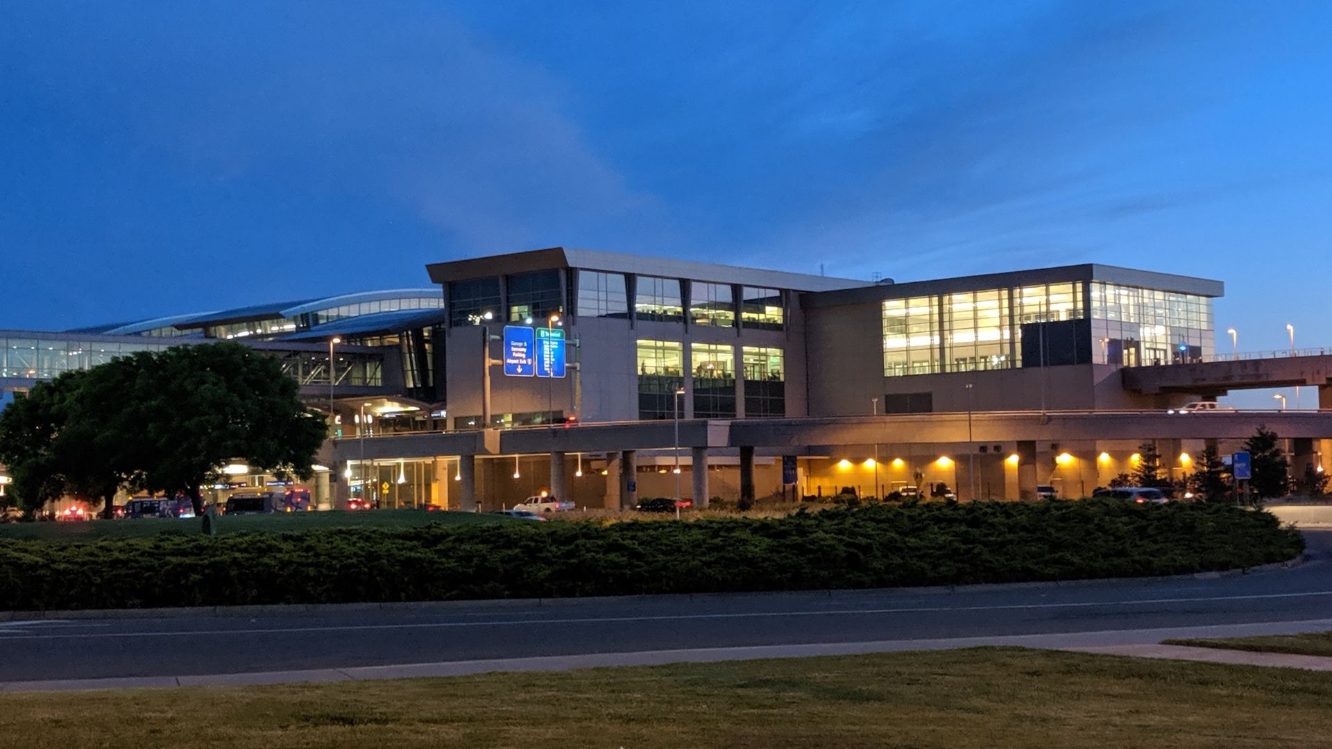 A wide exterior shot of the Sacramento International Airport terminal building at dusk, with lights illuminating the structure and surrounding landscape, including a grassy median and roadway in the foreground.