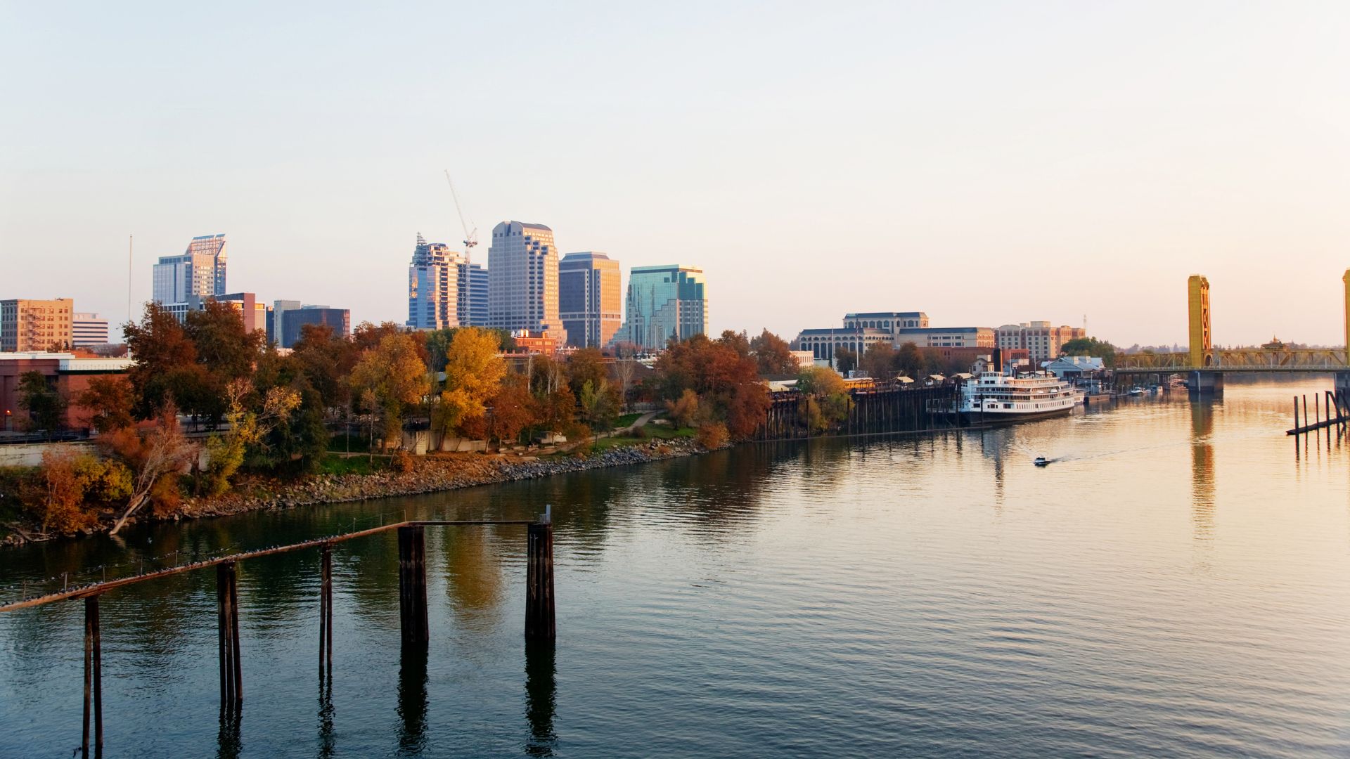 A panoramic view of the Sacramento, California skyline at sunset, featuring the Sacramento River in the foreground reflecting the warm light, the iconic golden Tower Bridge spanning the river on the right, and the city's modern high-rise buildings visible in the distance on the left bank, surrounded by autumn-colored trees.