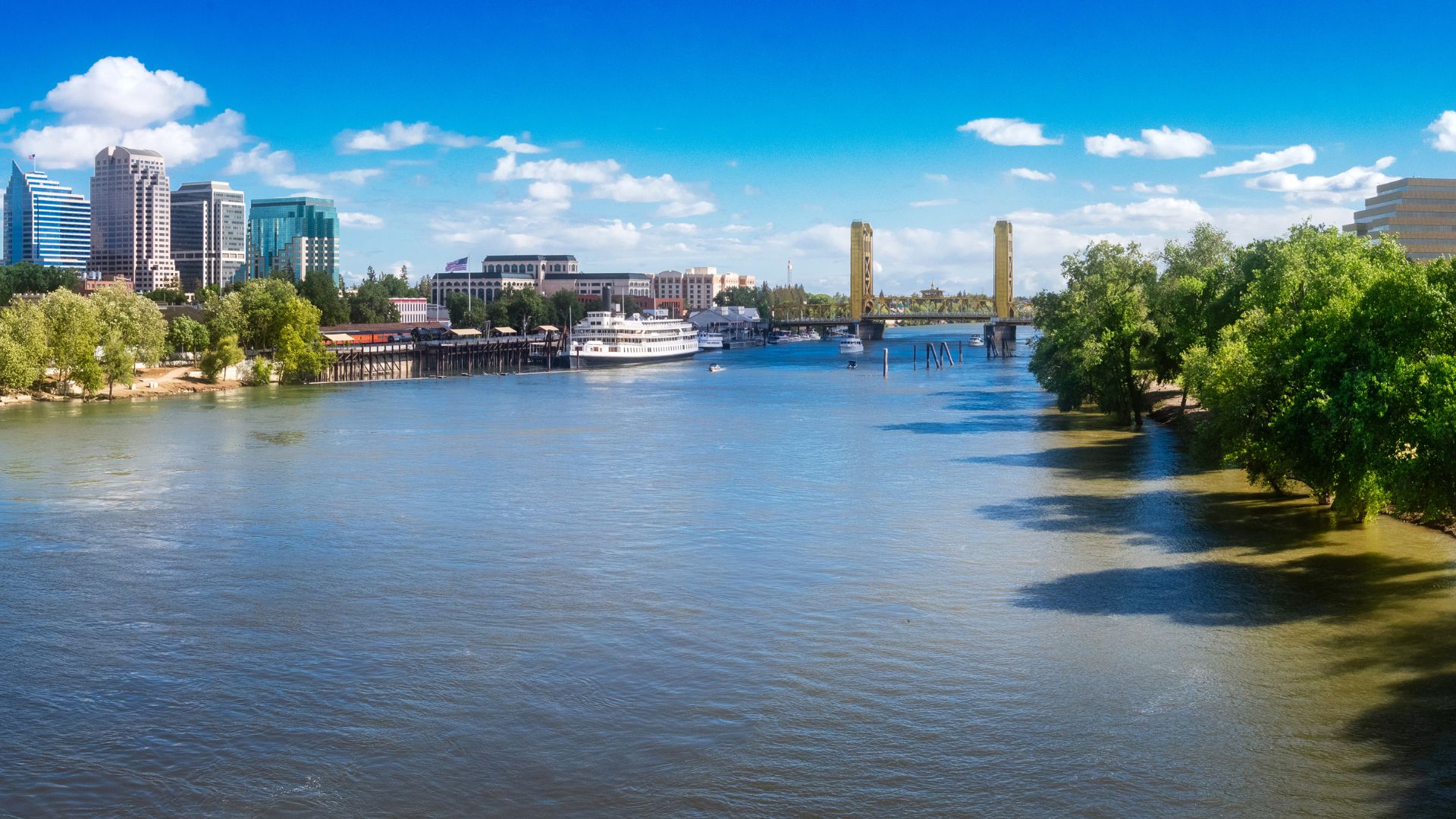 A wide shot of the Sacramento River in California, featuring the distinctive golden Tower Bridge on the right and the city skyline with tall buildings on the left bank, all under a bright blue sky with scattered white clouds. A large white boat is docked near the left bank, and lush green trees line both sides of the river.