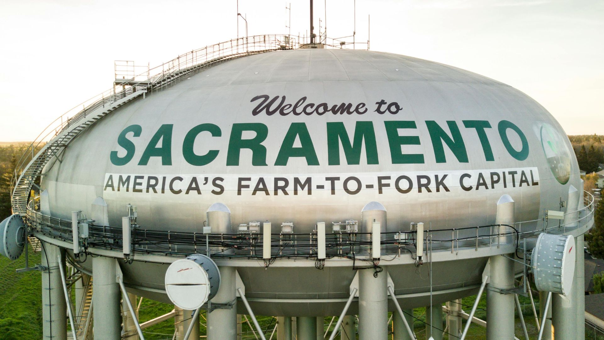 A large, light-colored water tower with "Welcome to SACRAMENTO AMERICA'S FARM-TO-FORK CAPITAL" written in green letters across its spherical tank, set against a clear sky with a hint of sunset. The tower is supported by a tall metal structure, and various antennas and equipment are visible on its surface.