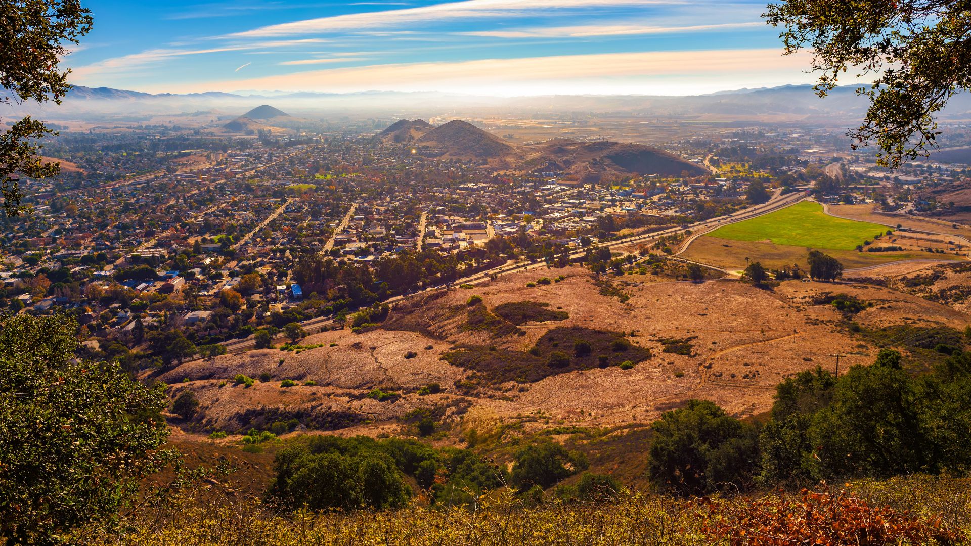 A high-angle panoramic view of San Luis Obispo, California, showing the city nestled among hills and fields under a bright, partly cloudy sky, with Cerro San Luis Obispo visible.