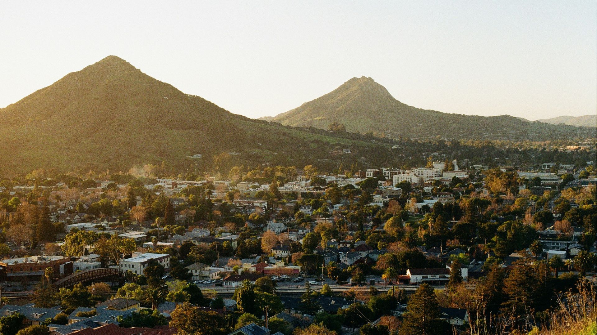 An aerial view of San Luis Obispo, California, showing a sprawling city surrounded by prominent, sunlit hills and mountains.
