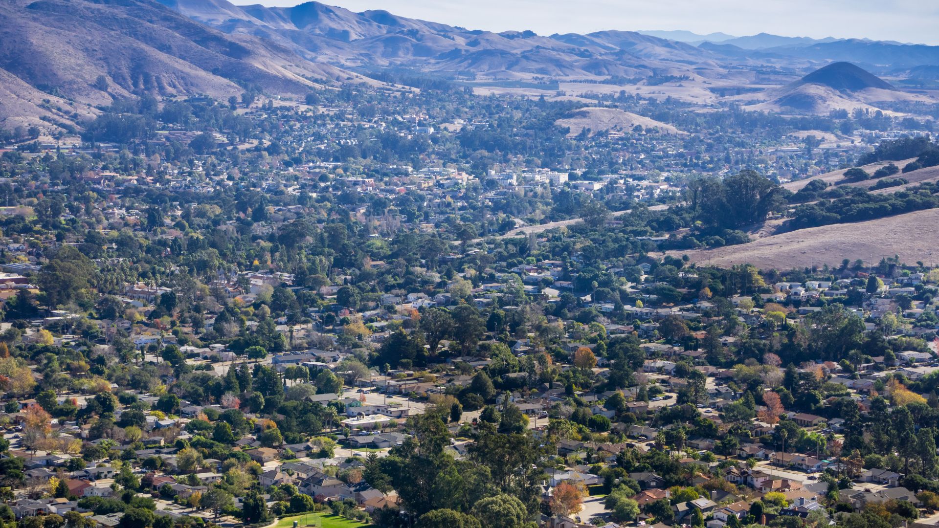 A panoramic view of San Luis Obispo, California, showing a sprawling city with numerous buildings and trees nestled within a valley, surrounded by rolling hills and mountains under a clear sky. 