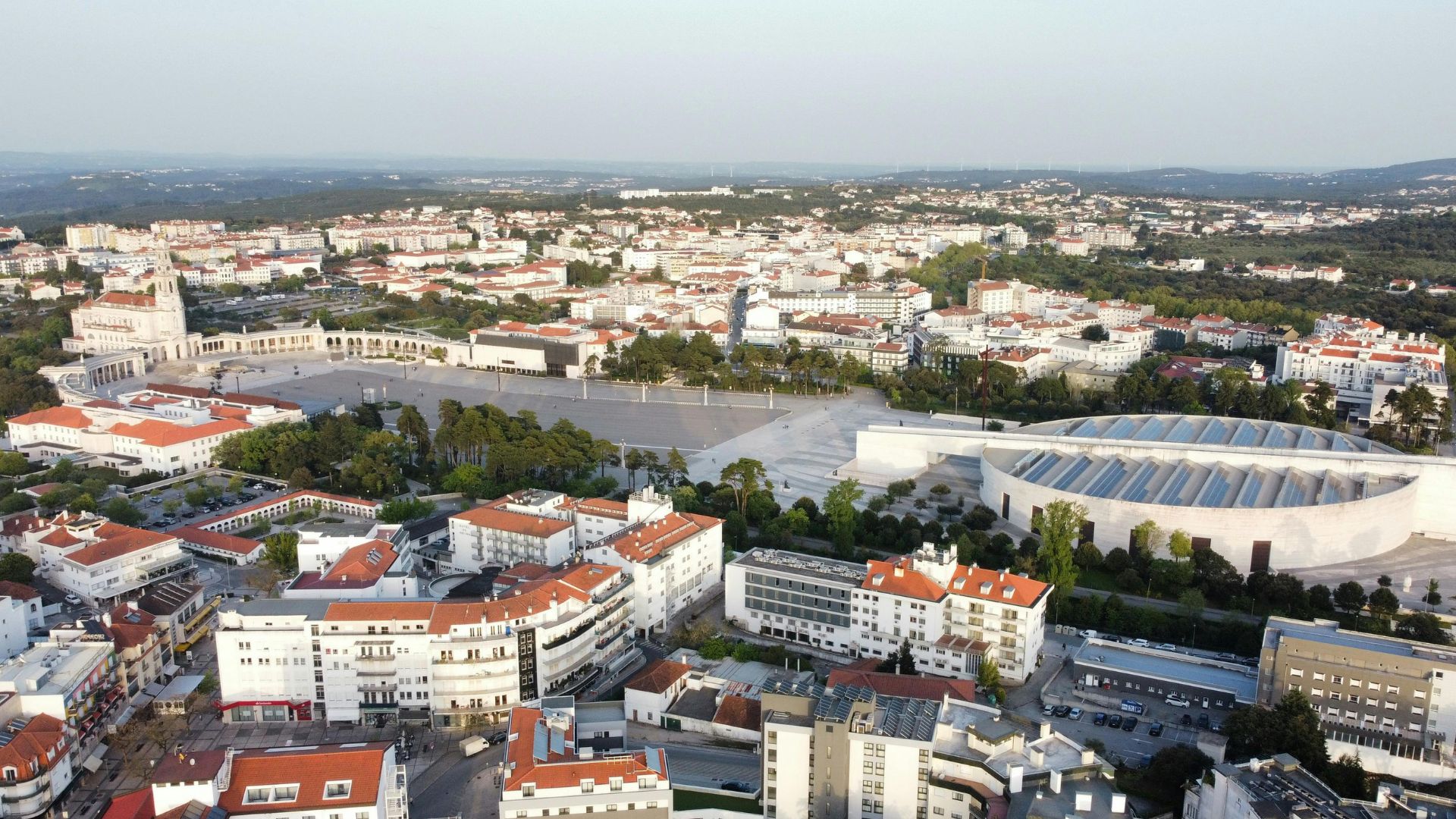 An aerial view of the Sanctuary of Our Lady of Fátima in central Portugal, showcasing the vast esplanade, the modern Basilica of the Holy Trinity, and the surrounding urban area of Fátima under a clear sky.