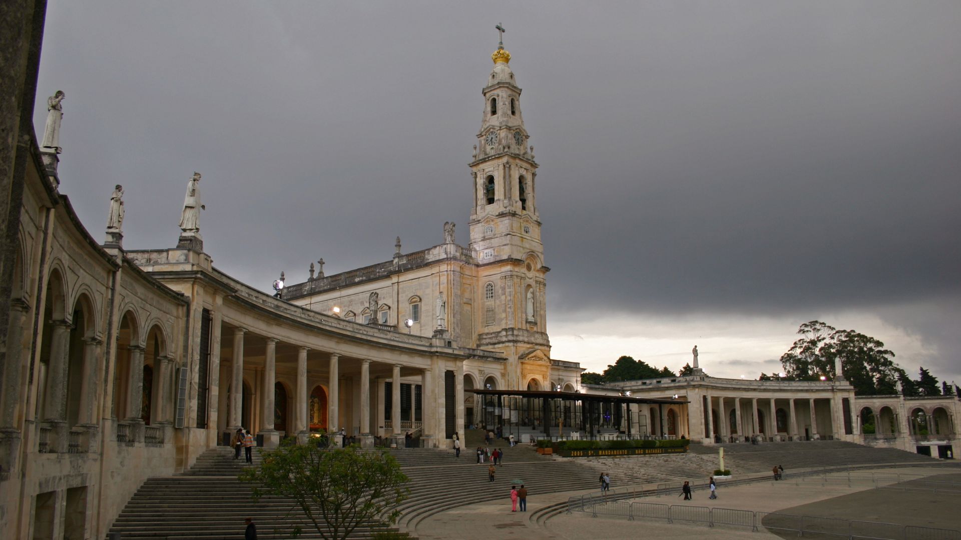 A wide shot of the Sanctuary of Our Lady of Fátima in central Portugal, featuring the prominent white Basilica of Our Lady of the Rosary with its tall bell tower, set against a bright blue sky with scattered white clouds, and surrounded by colonnades on either side. 