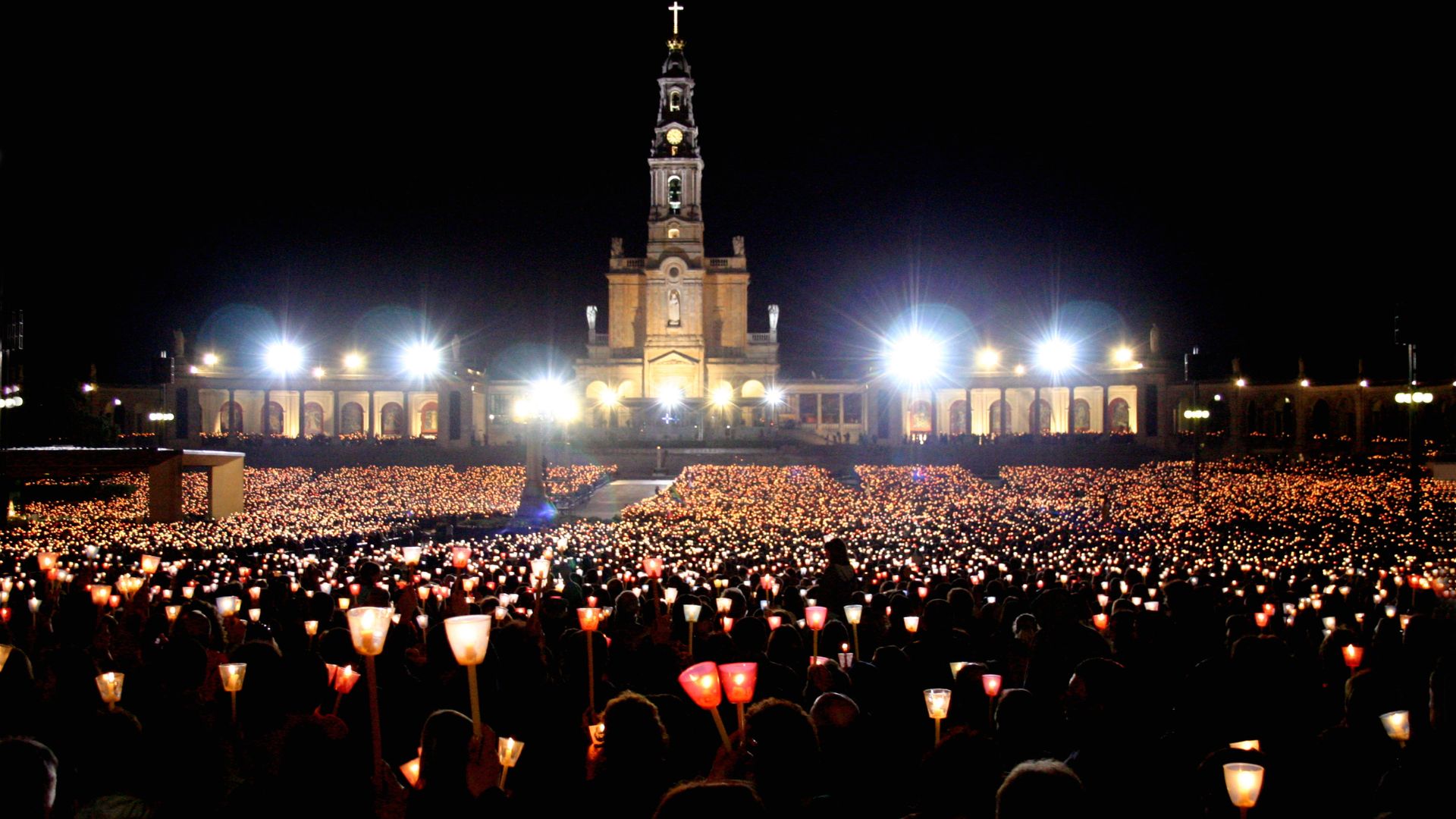 A nighttime image of the Santuário de Fátima in central Portugal, featuring the illuminated Basilica of Our Lady of the Rosary with its prominent clock tower and cross, overlooking a vast plaza filled with a dense crowd of pilgrims holding lit candles. The scene conveys a sense of spiritual gathering and devotion. 