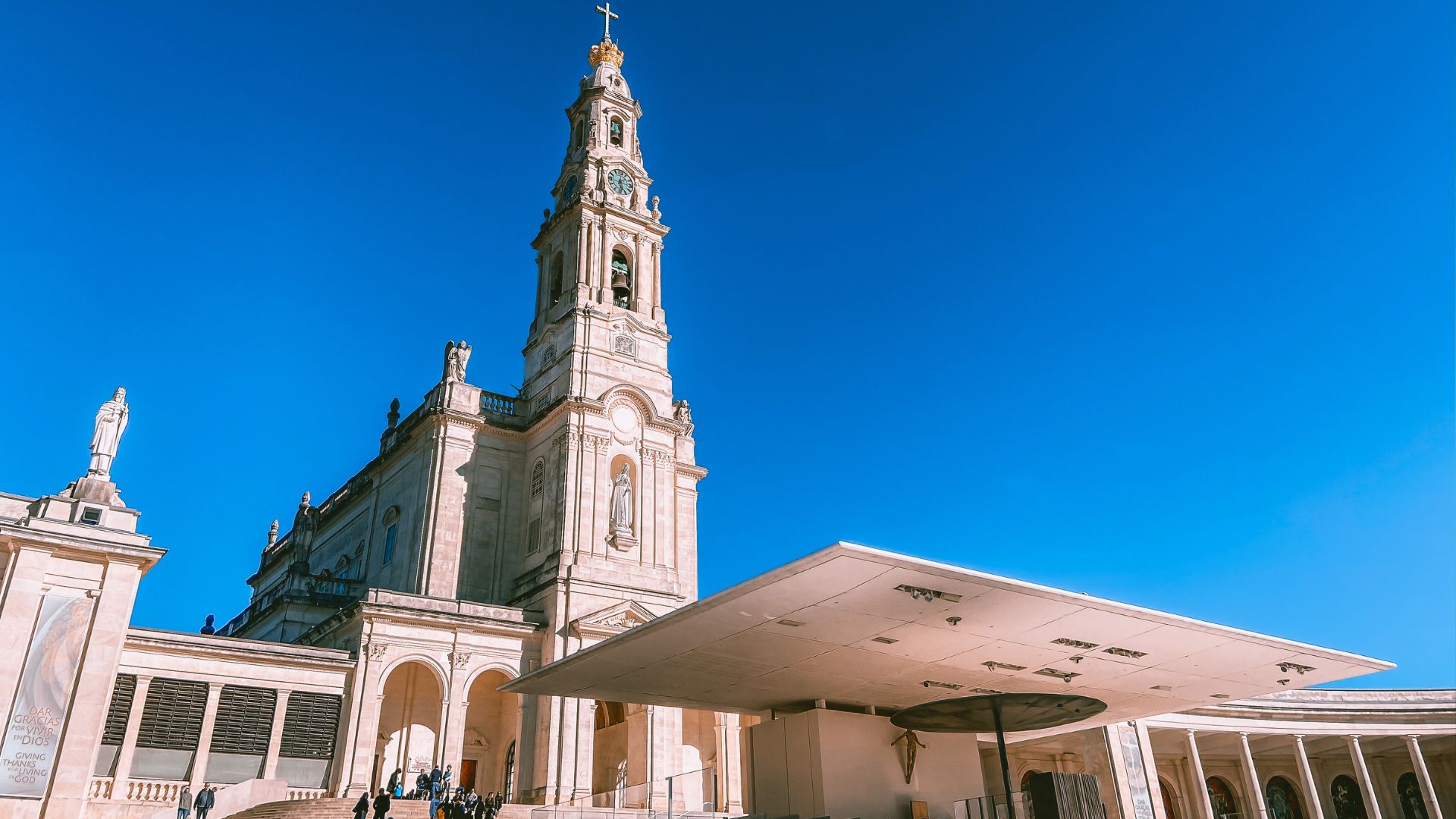 A wide shot of the Sanctuary of Our Lady of Fátima in central Portugal, featuring the prominent white Basilica of Our Lady of the Rosary with its tall bell tower, set surrounded by colonnades on either side. 
