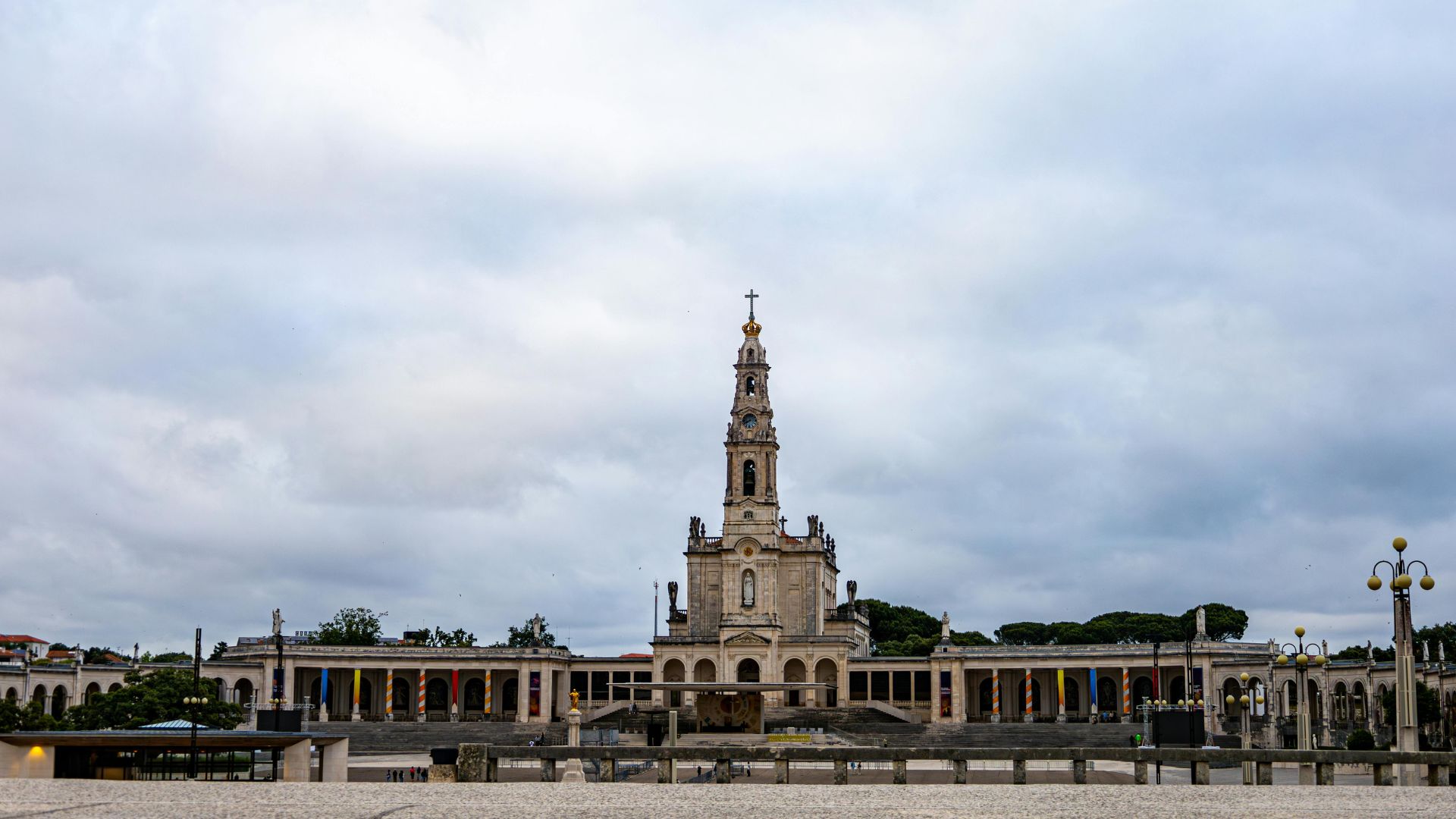 A wide shot of the Sanctuary of Our Lady of Fátima in central Portugal, featuring the prominent white Basilica of Our Lady of the Rosary with its tall bell tower under a bright blue sky with scattered clouds. The foreground 