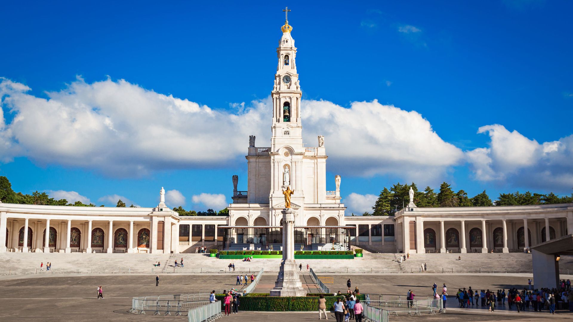A wide shot of the Sanctuary of Our Lady of Fátima in central Portugal, featuring the prominent white Basilica of Our Lady of the Rosary with its tall bell tower under a bright blue sky with scattered clouds. The foreground 