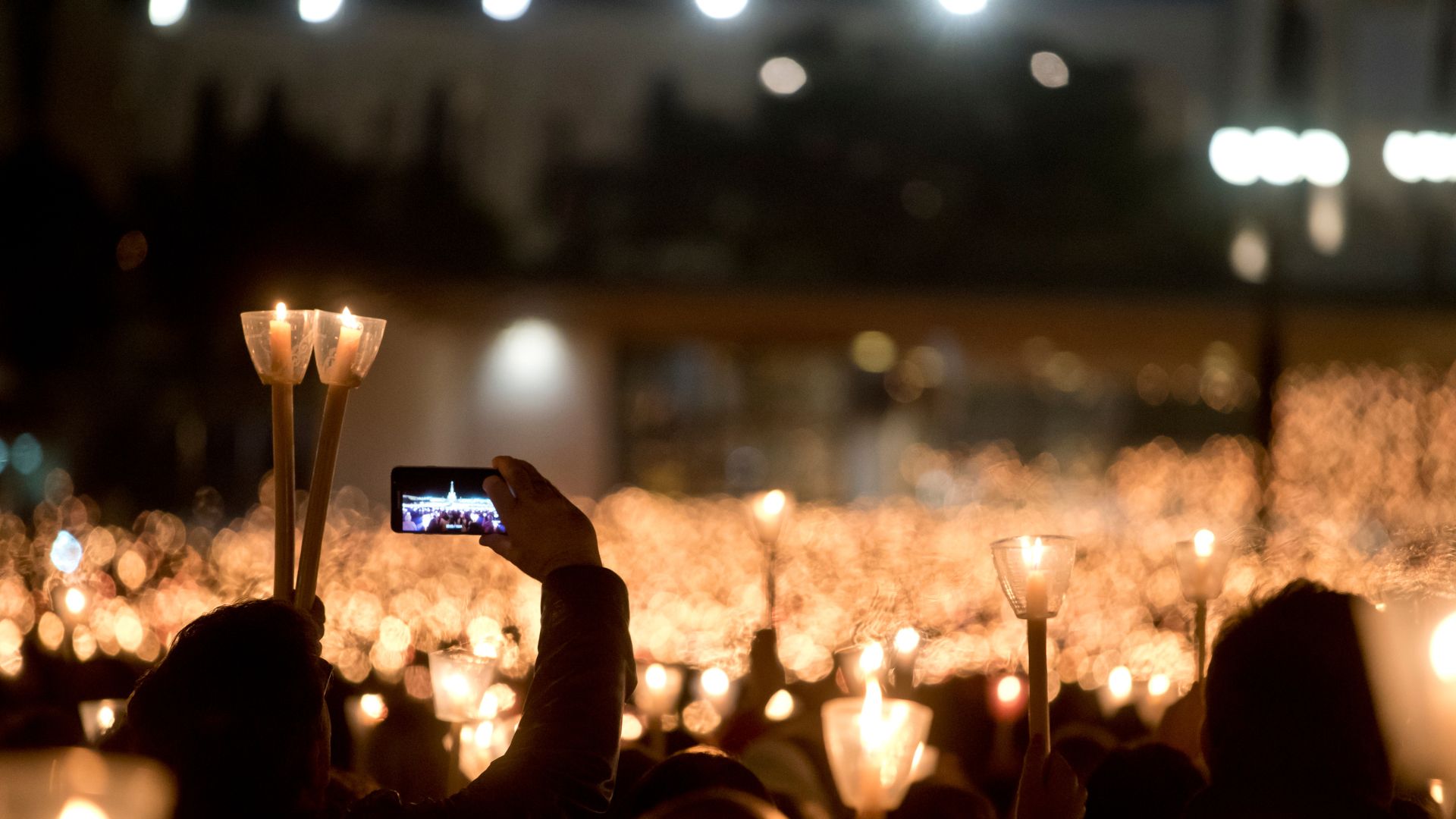 A nighttime image of the Santuário de Fátima in central Portugal, featuring the illuminated Basilica of Our Lady of the Rosary with its prominent clock tower and cross, overlooking a vast plaza filled with a dense crowd of pilgrims holding lit candles. The scene conveys a sense of spiritual gathering and devotion. 
