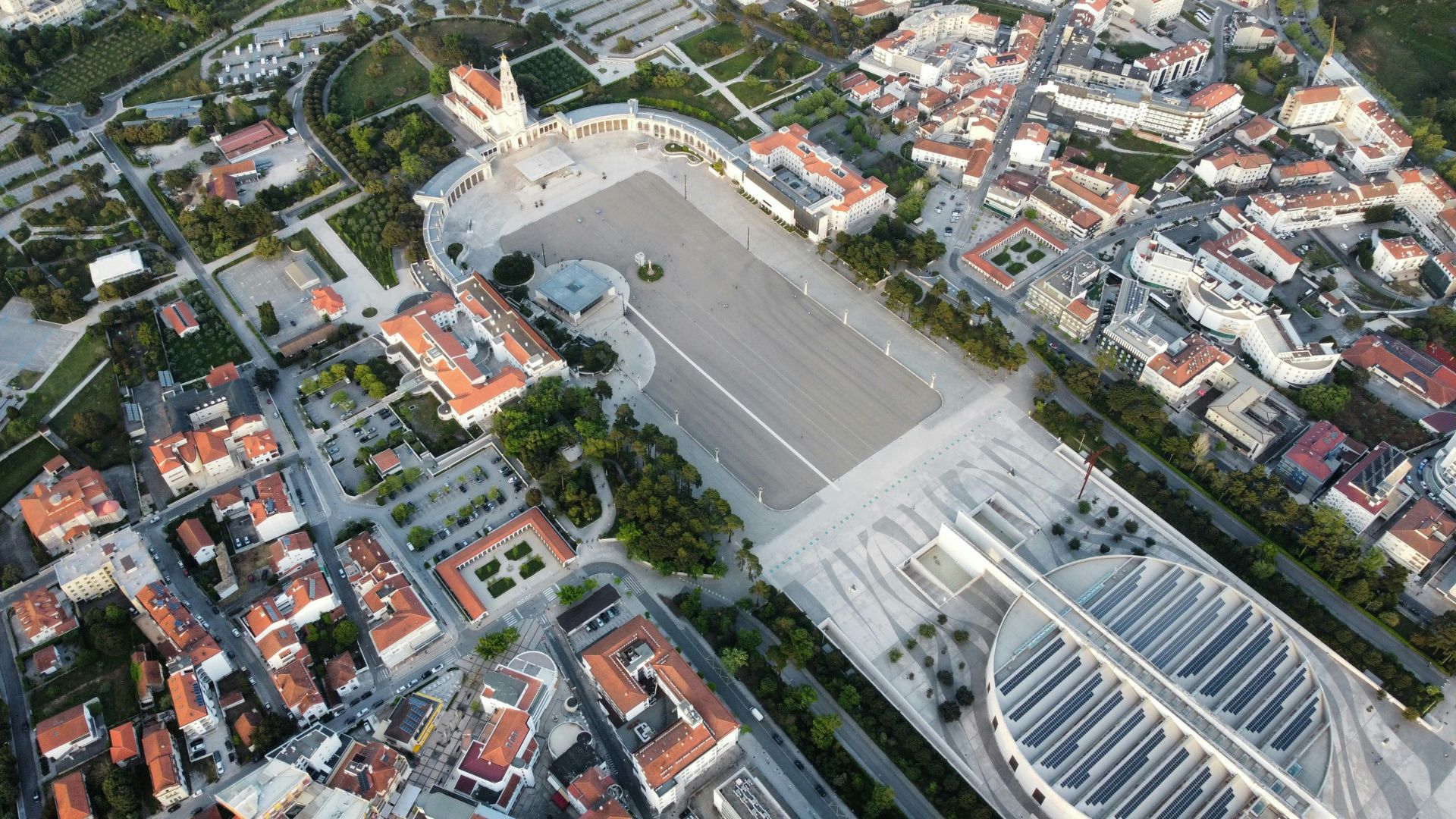 An aerial view of the Sanctuary of Fátima in Portugal, showcasing the vast esplanade, the Basilica of Our Lady of the Rosary, and the modern Basilica of the Most Holy Trinity, surrounded by the town's buildings. 