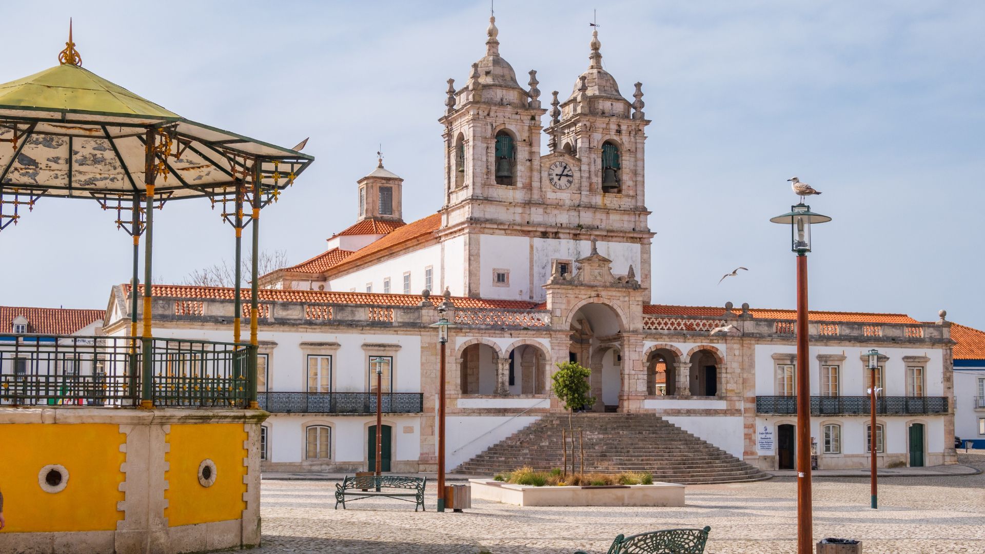 A wide shot of the Sanctuary of Our Lady of Nazaré, a large white church with two prominent bell towers, situated at the top of a grand staircase, with a traditional yellow and green bandstand in the foreground on the left.