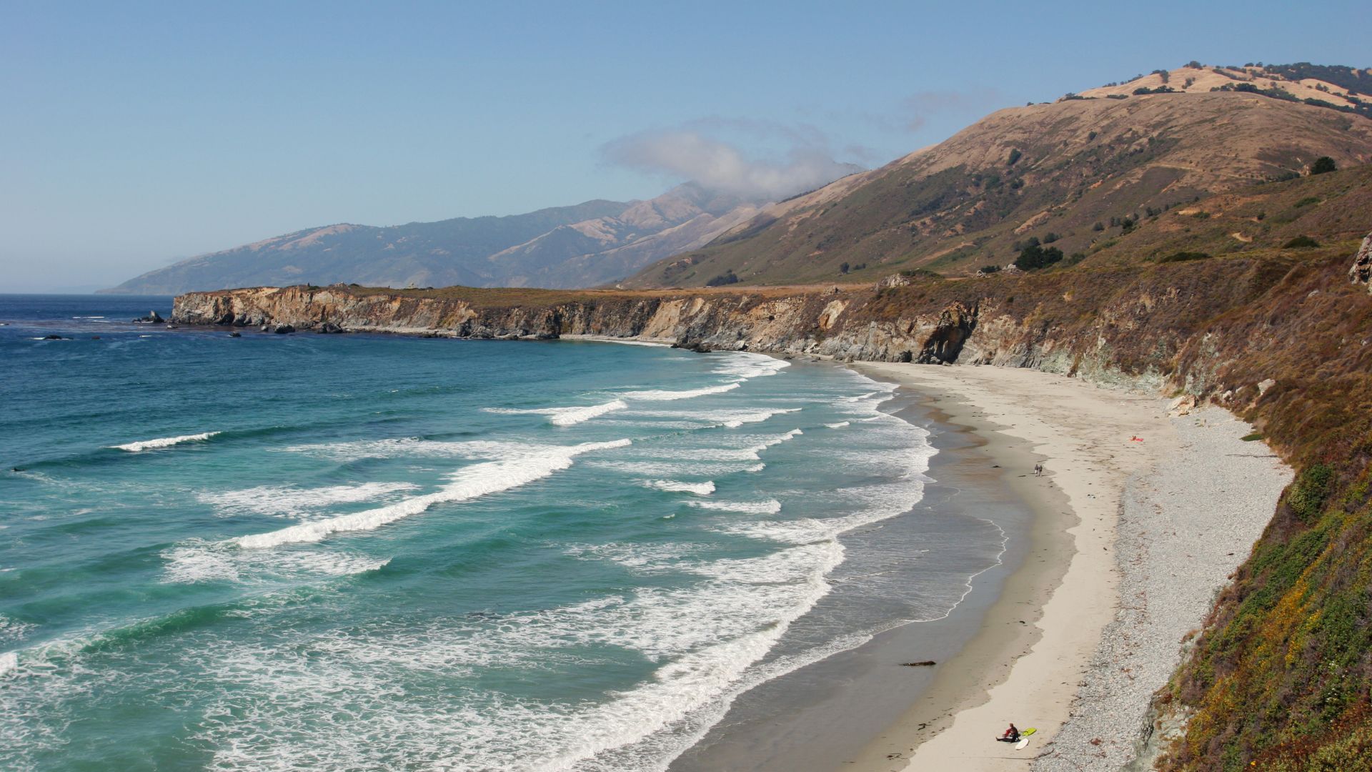 A wide, crescent-shaped beach with turquoise waves breaking on the shore, backed by a steep, rocky cliff and rolling hills under a clear blue sky. 