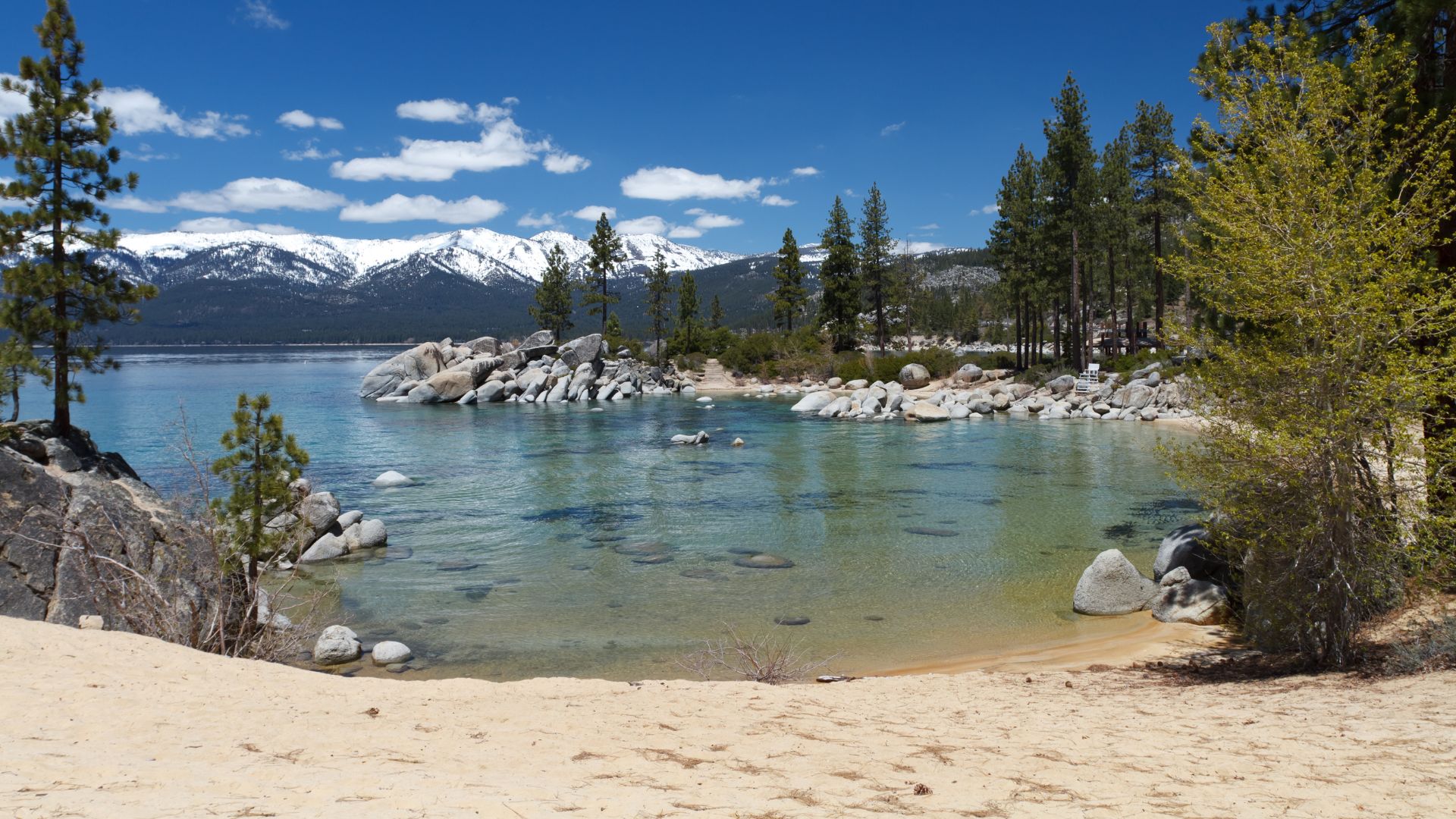 A serene panoramic view of Sand Harbor Beach at Lake Tahoe, featuring a sandy shore leading into clear, turquoise water with scattered boulders, framed by tall pine trees and snow-capped mountains under a bright blue sky with scattered clouds.