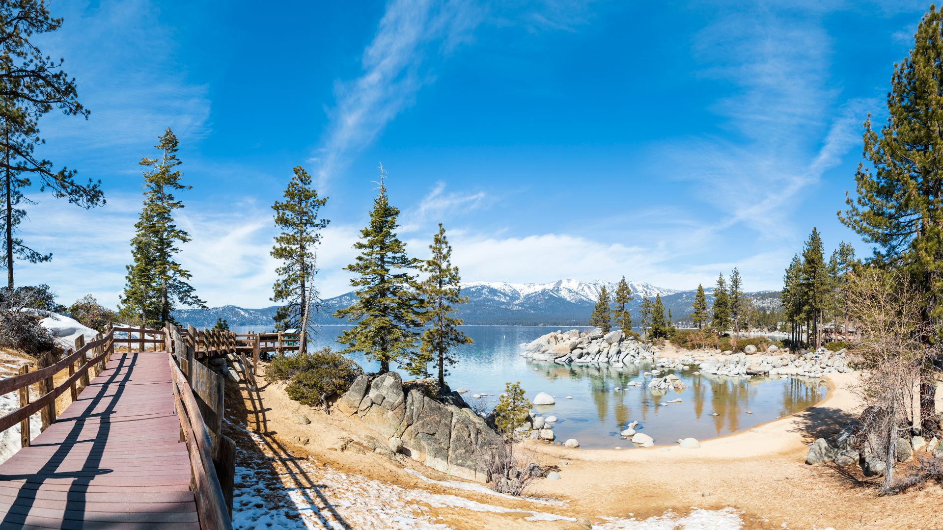 A panoramic view of Sand Harbor at Lake Tahoe, featuring a wooden boardwalk on the left, leading towards a sandy beach with clear blue water and scattered rocks. Pine trees line the shore, and snow-capped mountains rise in the distance under a bright blue sky with wispy clouds.