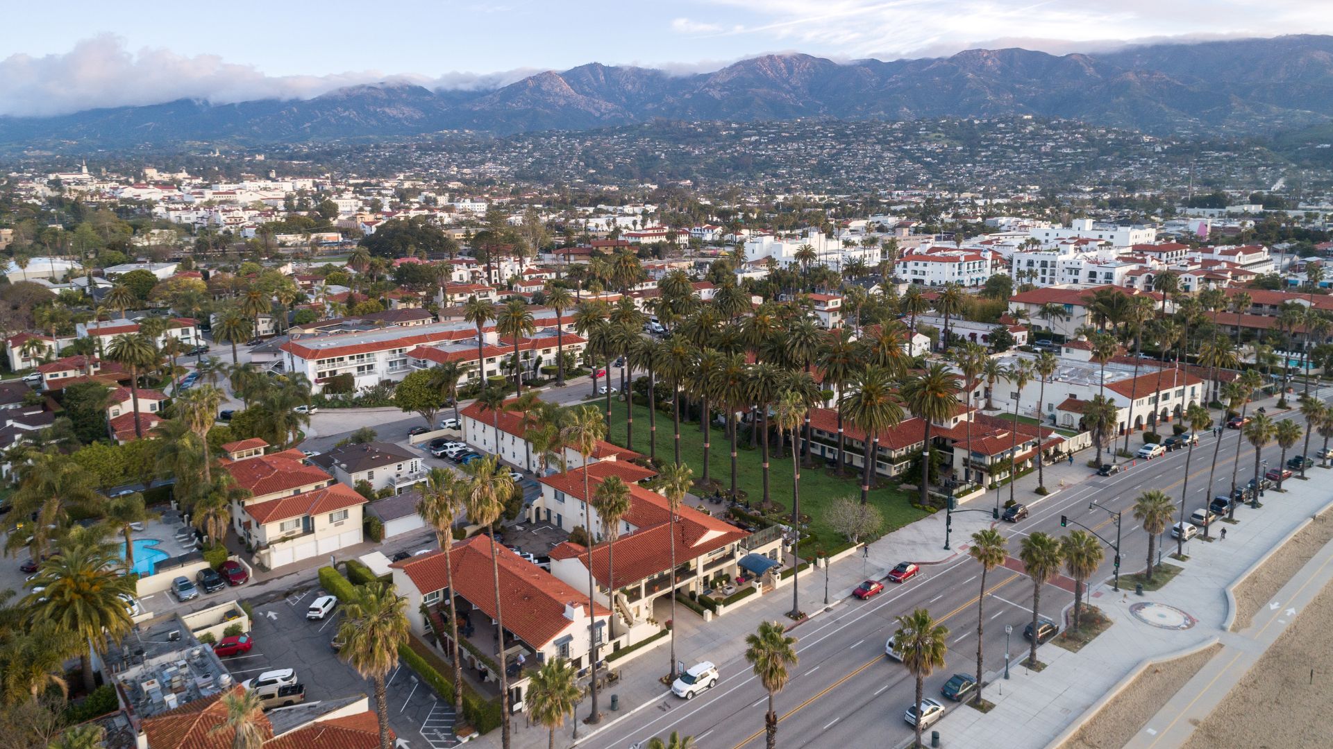 An aerial view of Santa Barbara, California, showcasing a coastal city with palm tree-lined streets, buildings featuring red-tiled roofs, and the Santa Ynez Mountains in the background. The Pacific Ocean is visible along the right side of the image.