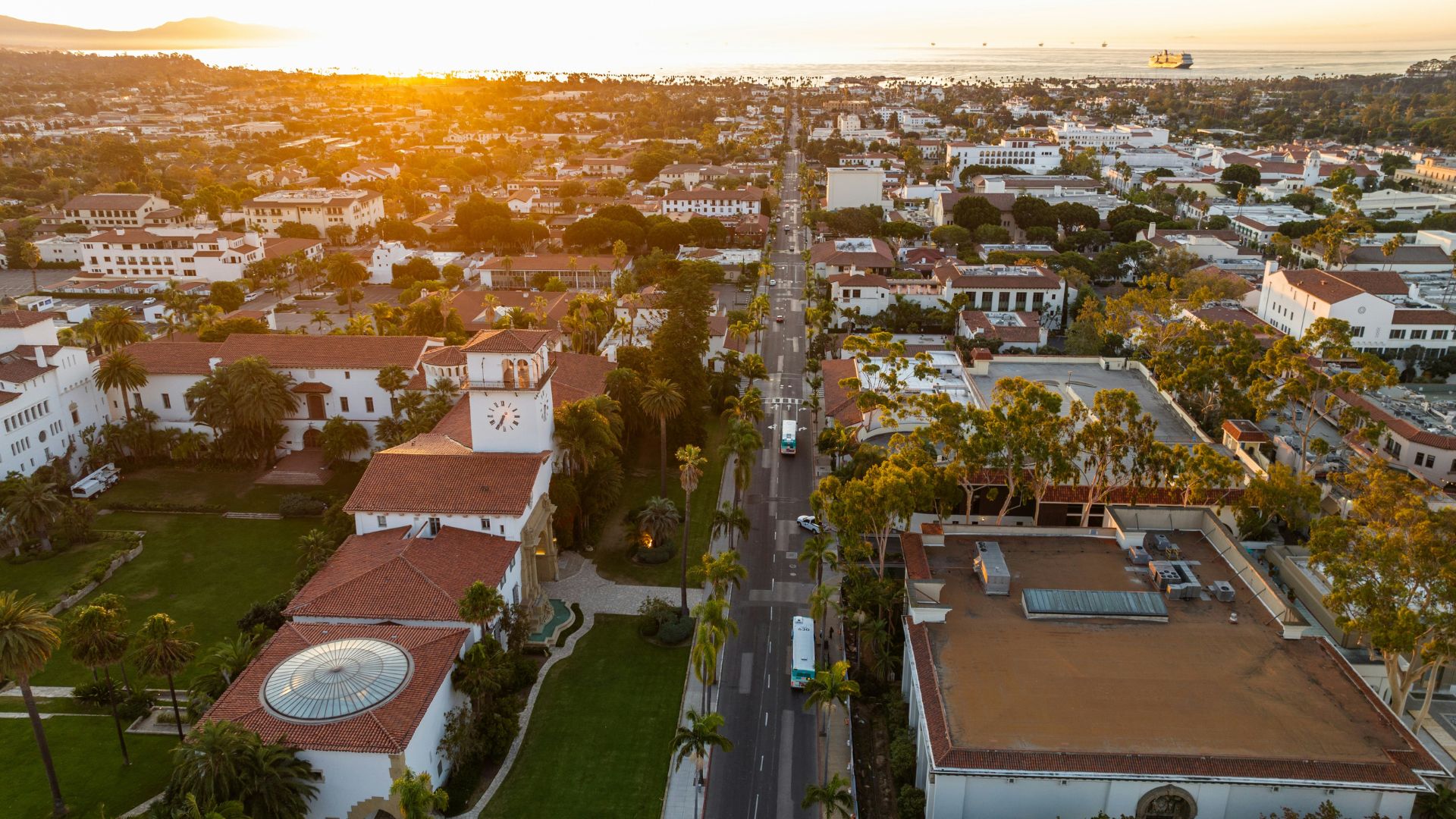 An aerial view of the Santa Barbara County Courthouse at sunset, showcasing its Spanish Colonial Revival architecture, including the distinctive clock tower and red-tiled roofs, surrounded by lush green grounds and a tree-lined street leading towards the ocean in the distance. 