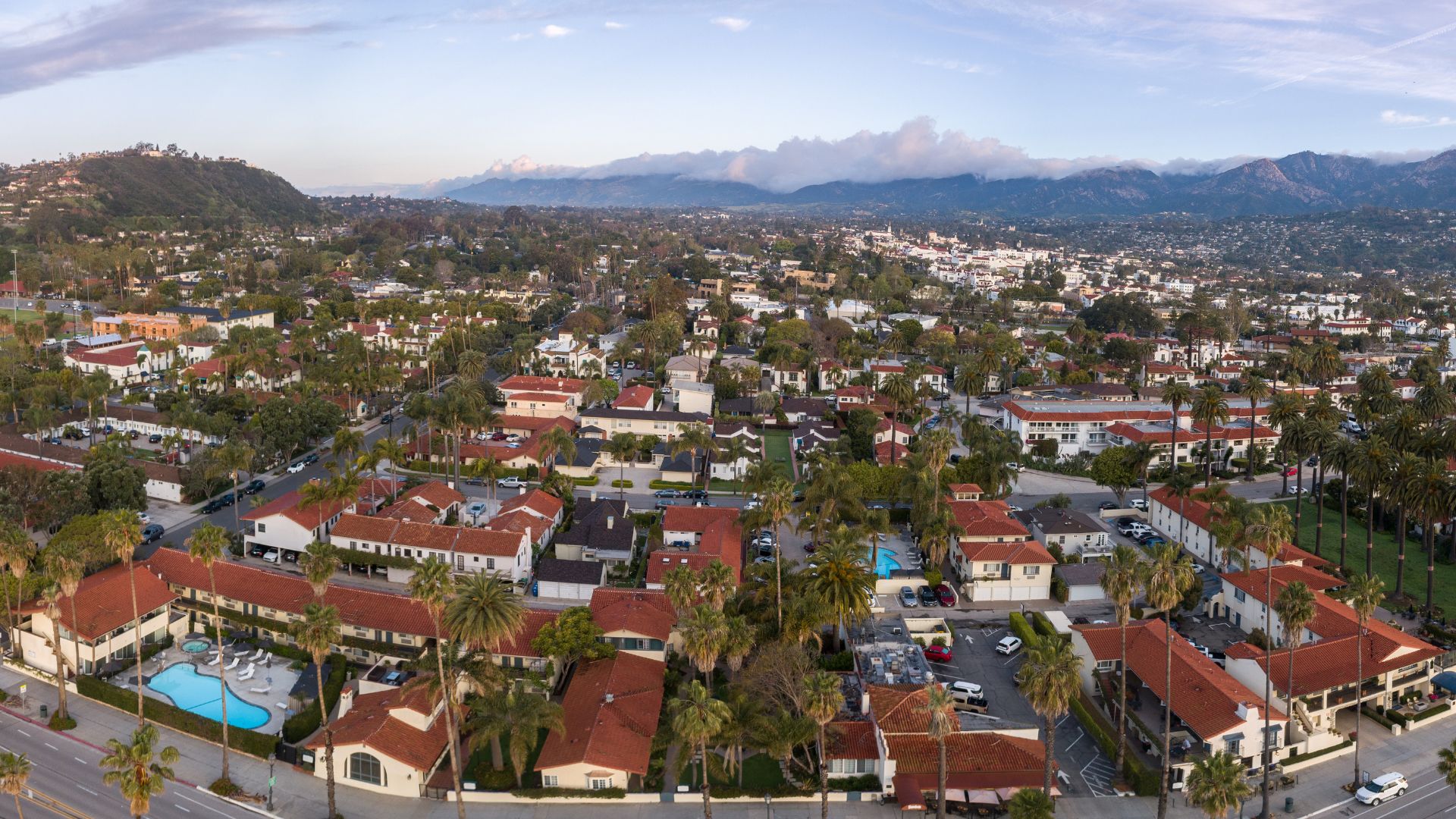 An aerial view of Santa Barbara, California, showcasing a coastal city with palm tree-lined streets, buildings featuring red-tiled roofs, and the Santa Ynez Mountains in the background. 