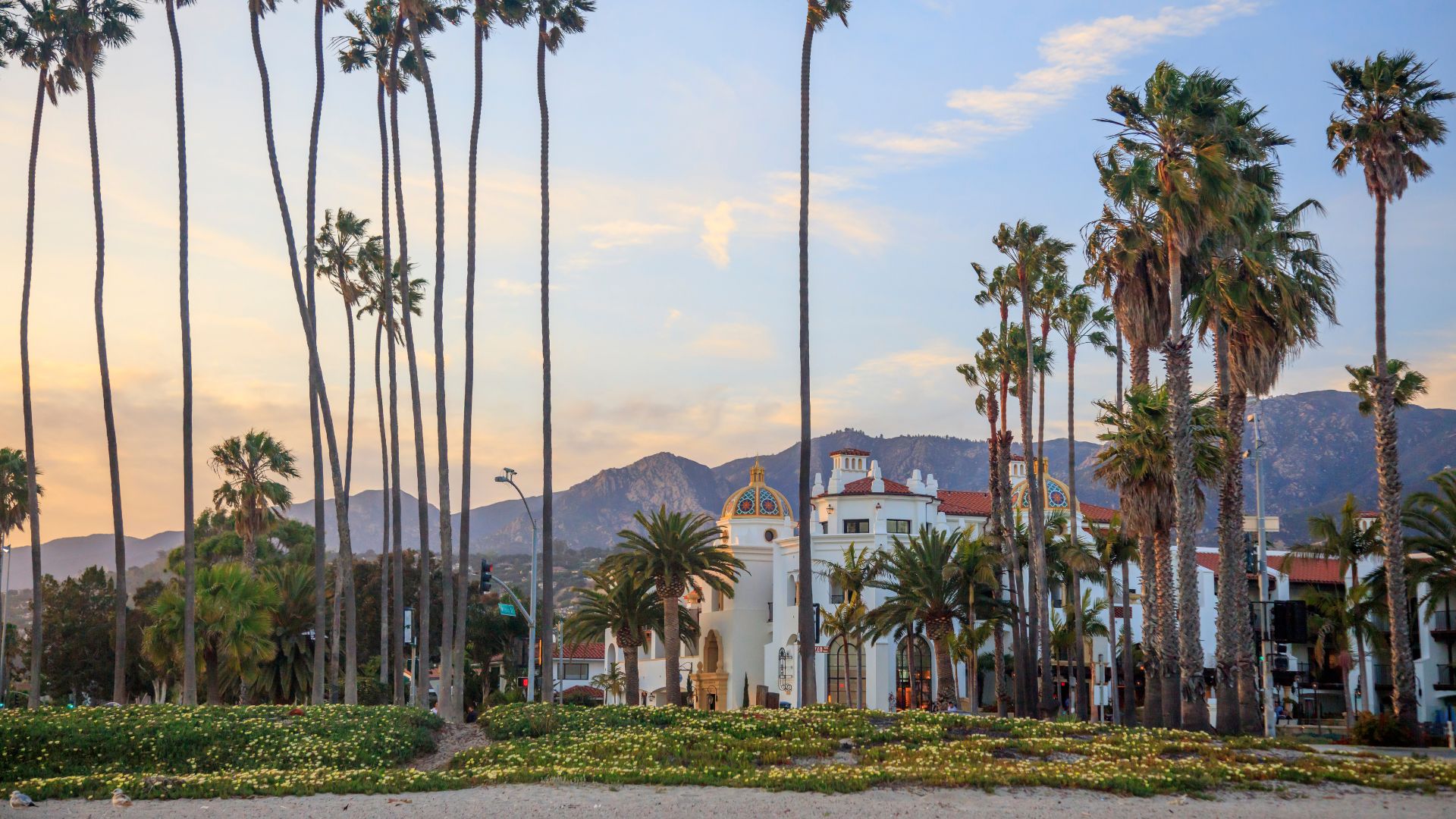 A scenic view of Santa Barbara, California, featuring tall palm trees lining a sandy beach with a large white building with a golden dome and red-tiled roof visible behind them, all set against a backdrop of mountains under a partly cloudy sky. 