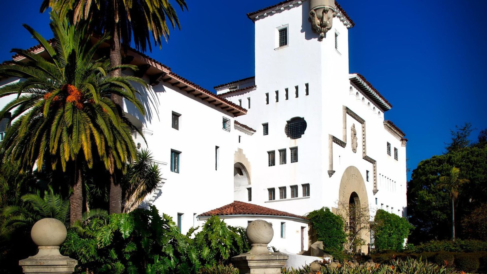 A striking image of the Santa Barbara County Courthouse, a Spanish Colonial Revival style building with a prominent tower, white stucco walls, and red tile roofs, set against a vibrant blue sky and surrounded by lush green gardens and palm trees.