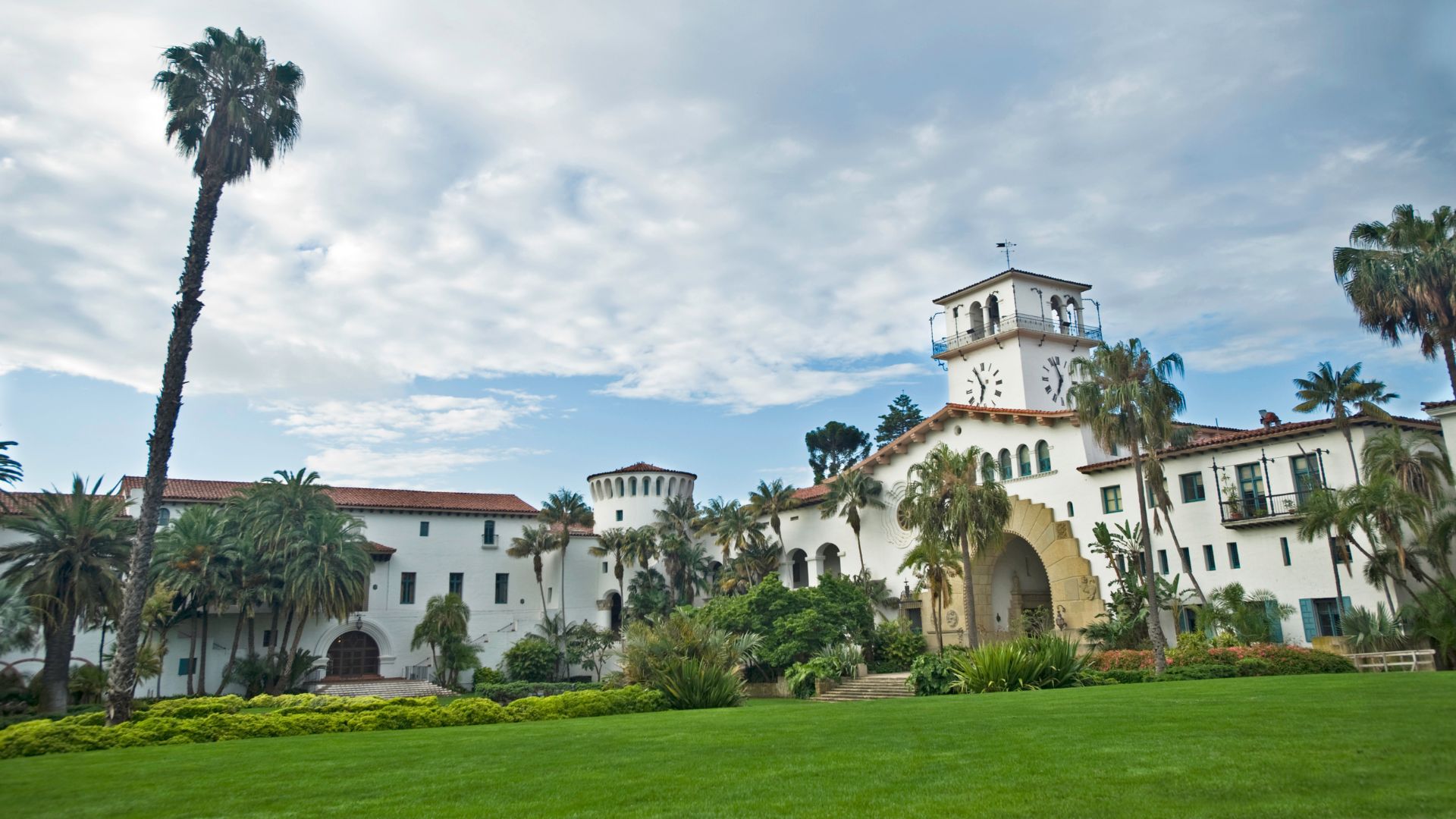 A wide-angle view of the Santa Barbara County Courthouse, a grand Spanish Colonial Revival building with a prominent clock tower, set against a partly cloudy sky. Lush green lawns and several tall palm trees are in the foreground and surround the building.