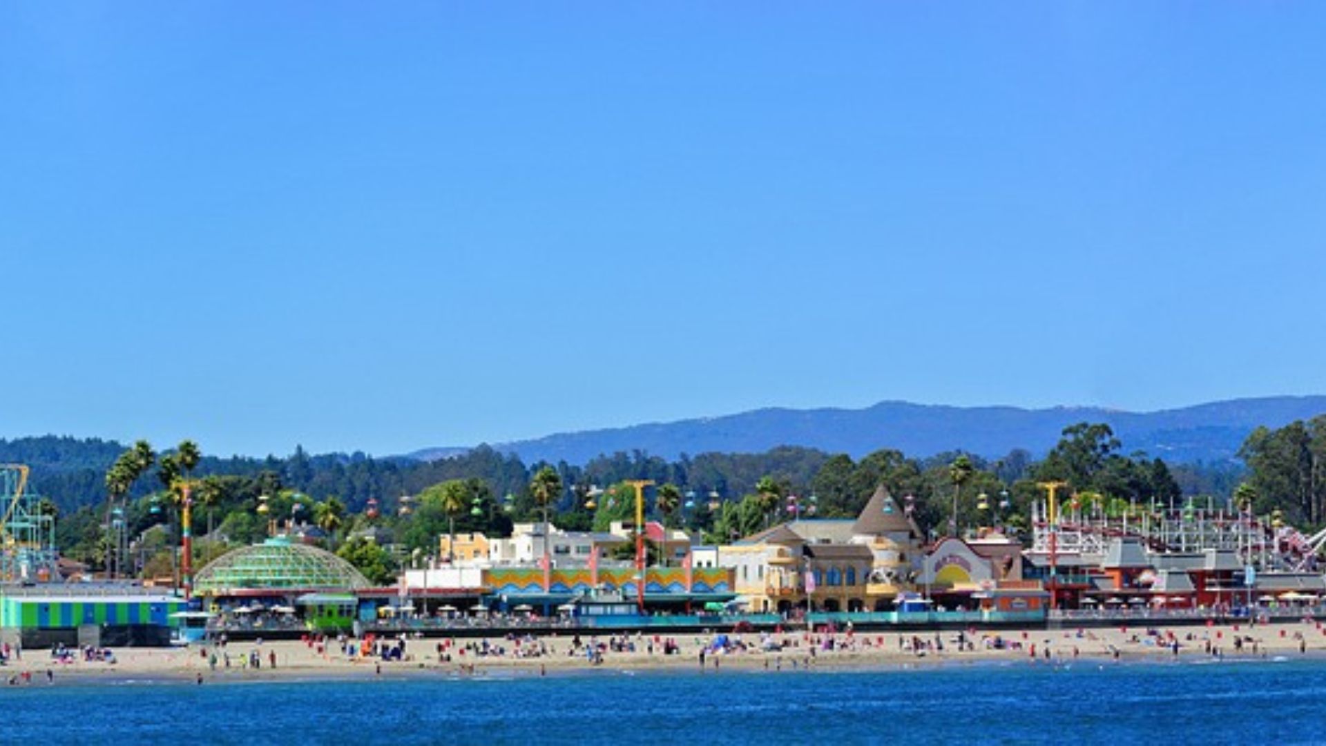 A panoramic view of the Santa Cruz Beach Boardwalk, showcasing a vibrant seaside amusement park with various rides, alongside a sandy beach with people, all set against a backdrop of green hills and a clear blue sky.