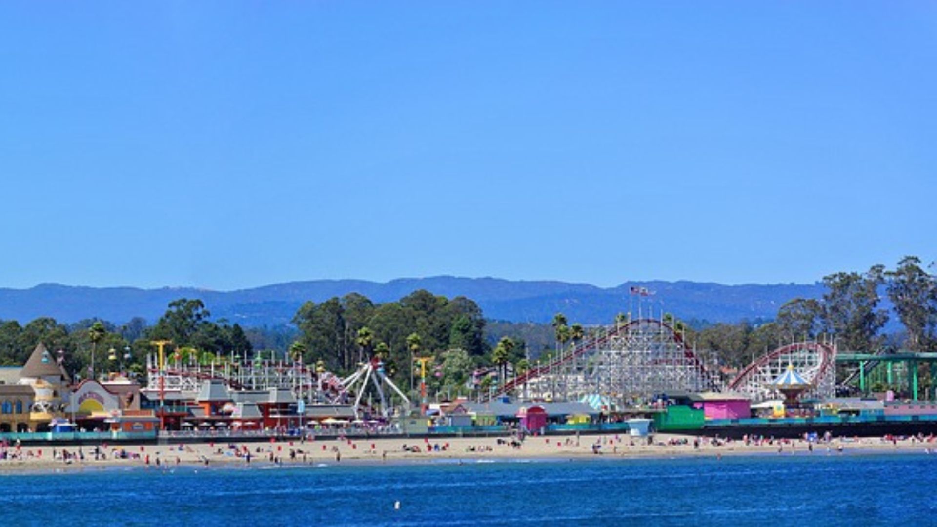 A panoramic view of the Santa Cruz Beach Boardwalk, showcasing a vibrant seaside amusement park with various rides, alongside a sandy beach with people, all set against a backdrop of green hills and a clear blue sky. 