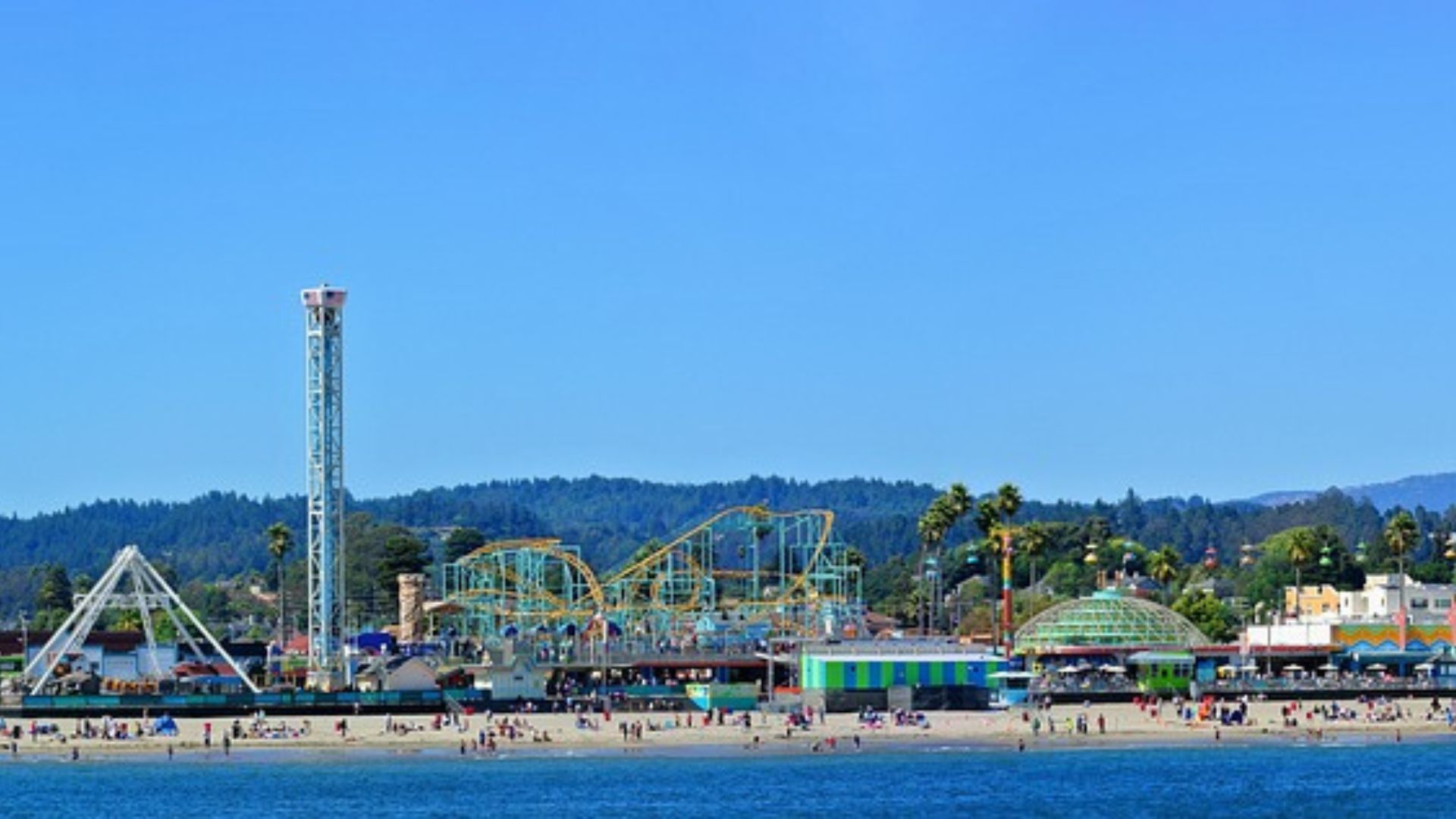 A panoramic view of the Santa Cruz Beach Boardwalk, showcasing a vibrant seaside amusement park with various rides, alongside a sandy beach with people, all set against a backdrop of green hills and a clear blue sky. 