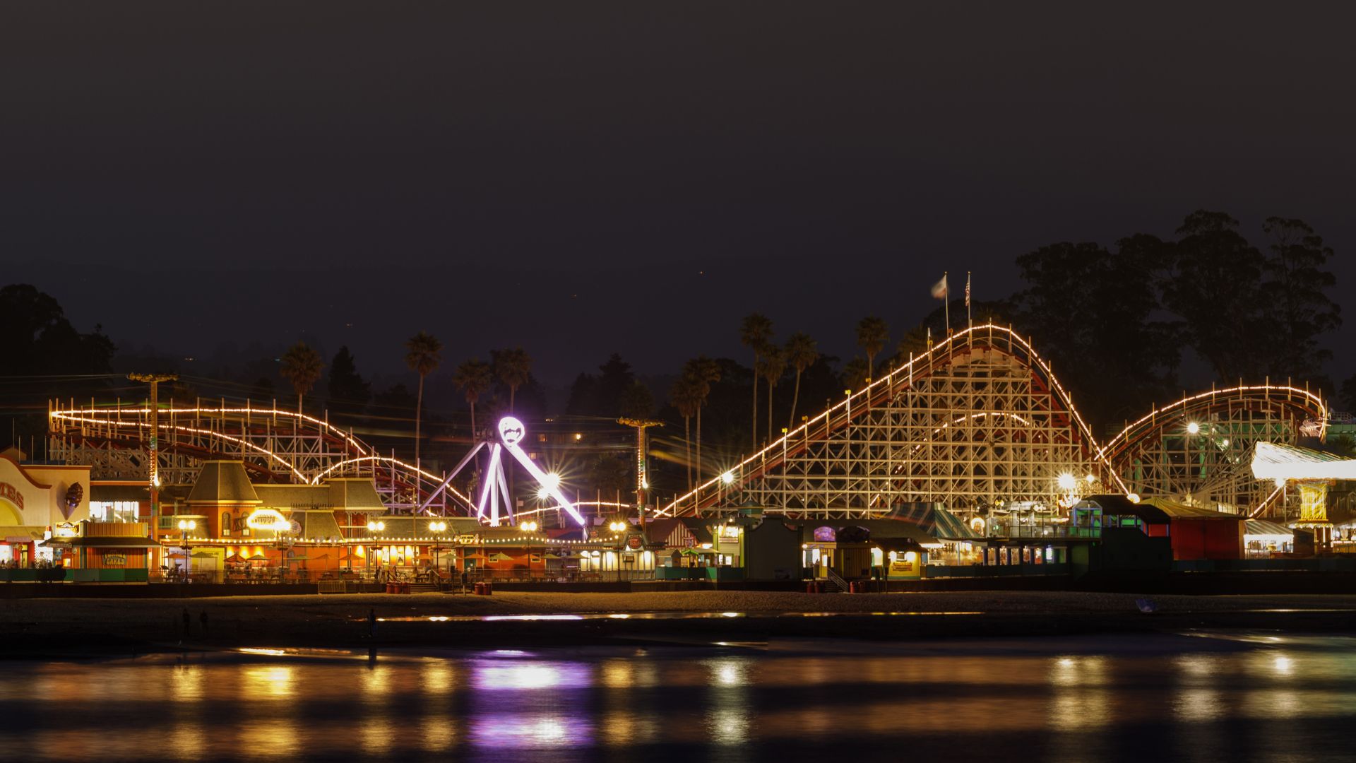 A nighttime long-exposure photograph captures the illuminated Santa Cruz Beach Boardwalk, featuring the iconic wooden structure of the Giant Dipper roller coaster and various amusement park rides and buildings lit up along the beachfront, with reflections shimmering on the water in the foreground.