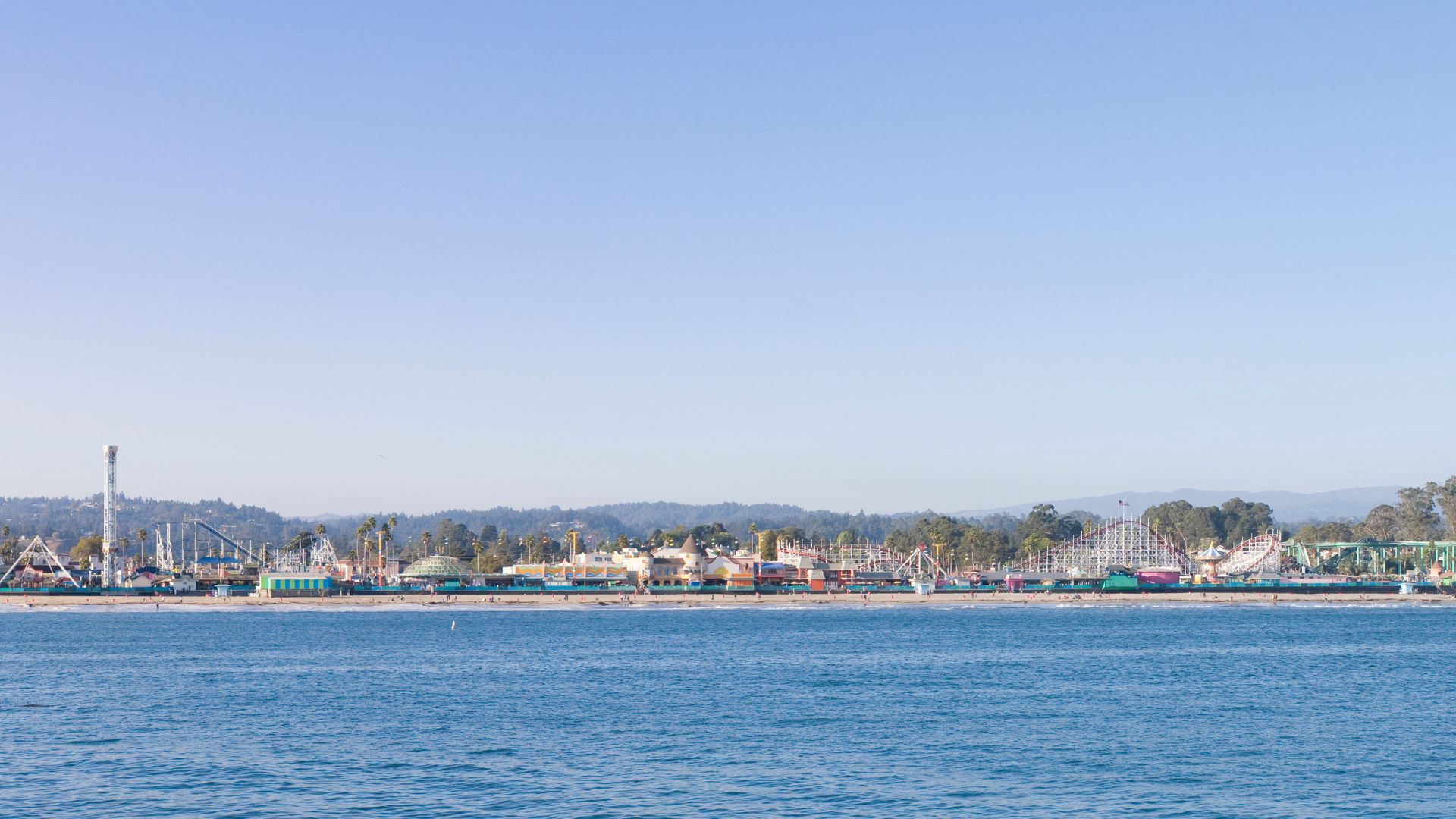 A panoramic view of the Santa Cruz Beach Boardwalk from across the water, showing the iconic Giant Dipper wooden roller coaster and other amusement park rides on the beach, with mountains visible in the background.