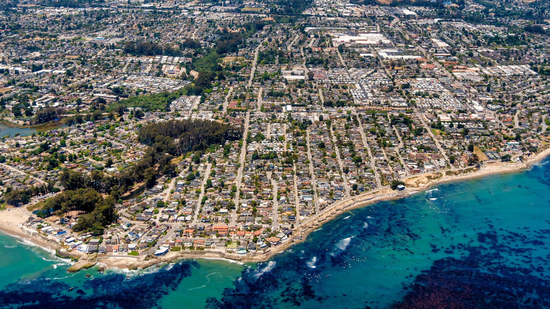 An aerial photograph showcases the city of Santa Cruz, California, stretching along a coastline with residential areas and green spaces extending inland from the ocean. The image captures the urban landscape transitioning into natural elements like beaches and the Pacific Ocean, with visible waves and underwater kelp beds near the shore.
