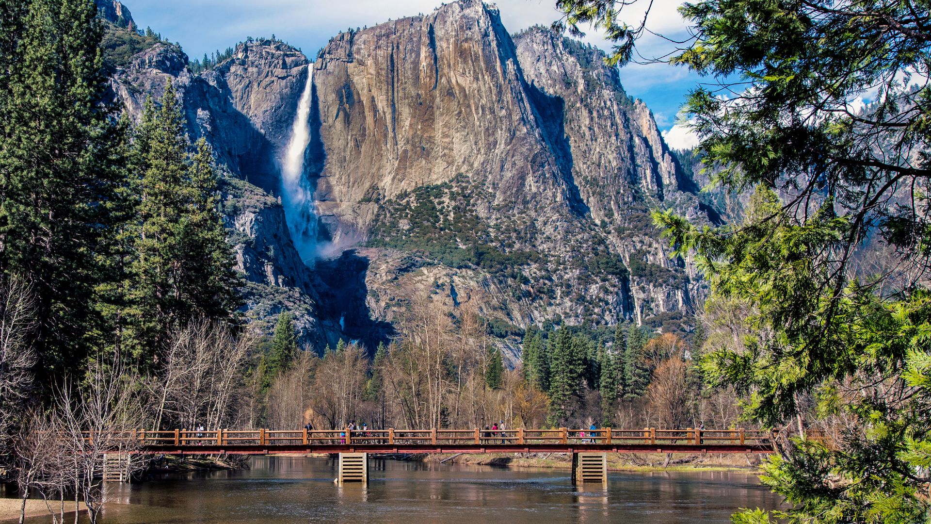 A scenic view of Sentinel Bridge over the Merced River in Yosemite National Park, with Yosemite Falls cascading down a granite cliff face in the background and surrounding evergreen trees.