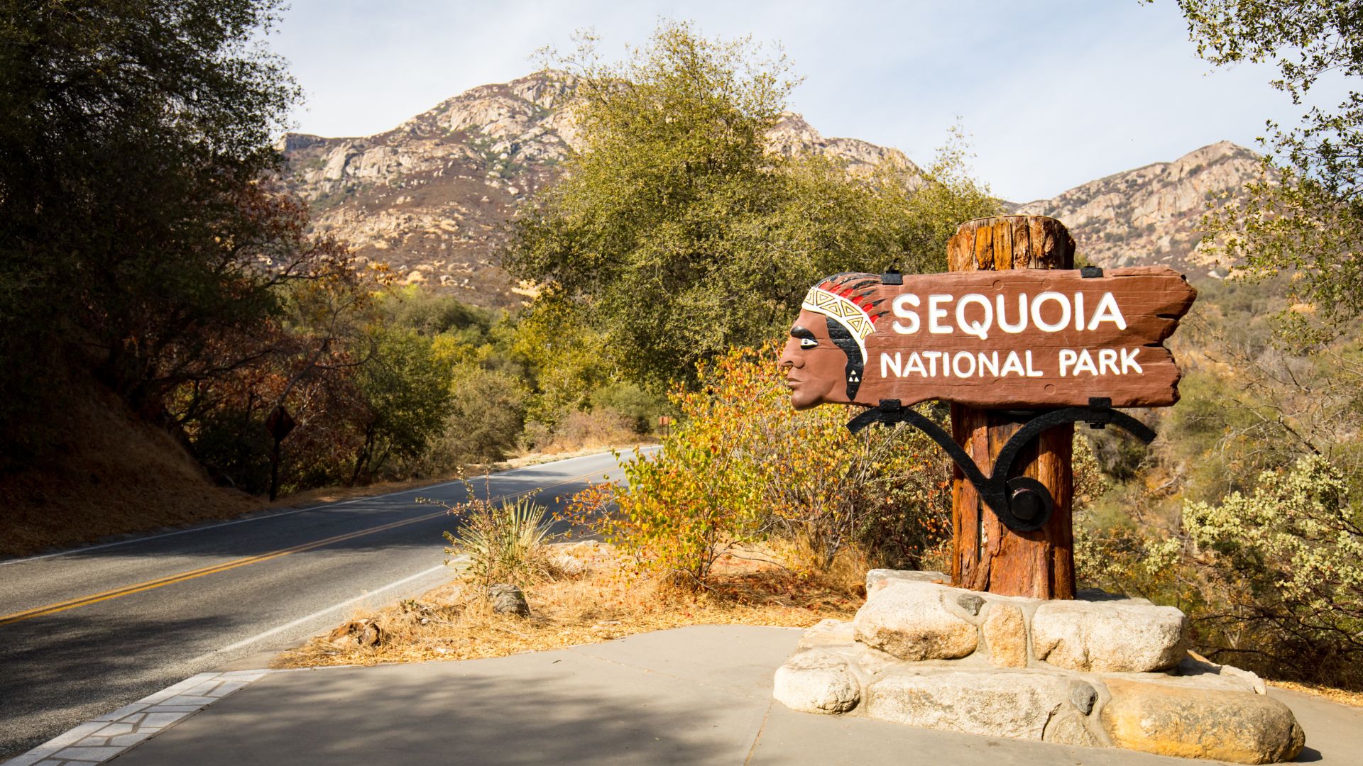 A wooden sign with "SEQUOIA NATIONAL PARK" carved into it stands beside a paved road, with a stylized depiction of a Native American profile on the left, set against a backdrop of trees and mountains under a bright sky.