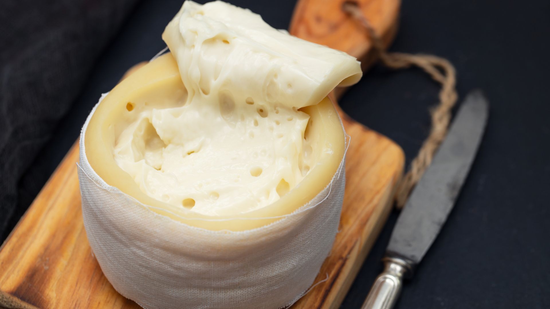 A round of soft, creamy Serra da Estrela cheese, partially unwrapped to reveal its pale yellow interior, sits on a wooden cutting board next to a knife and slices of bread, with a bottle of olive oil in the background.
