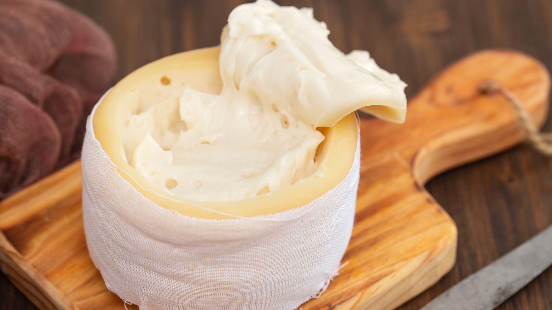A round of soft, creamy Serra da Estrela cheese, partially unwrapped to reveal its pale yellow interior, sits on a wooden cutting board next to a knife and slices of bread, with a bottle of olive oil in the background.