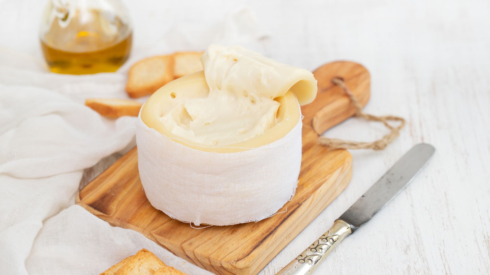 A round of soft, creamy Serra da Estrela cheese, partially unwrapped to reveal its pale yellow interior, sits on a wooden cutting board next to a knife and slices of bread, with a bottle of olive oil in the background.
