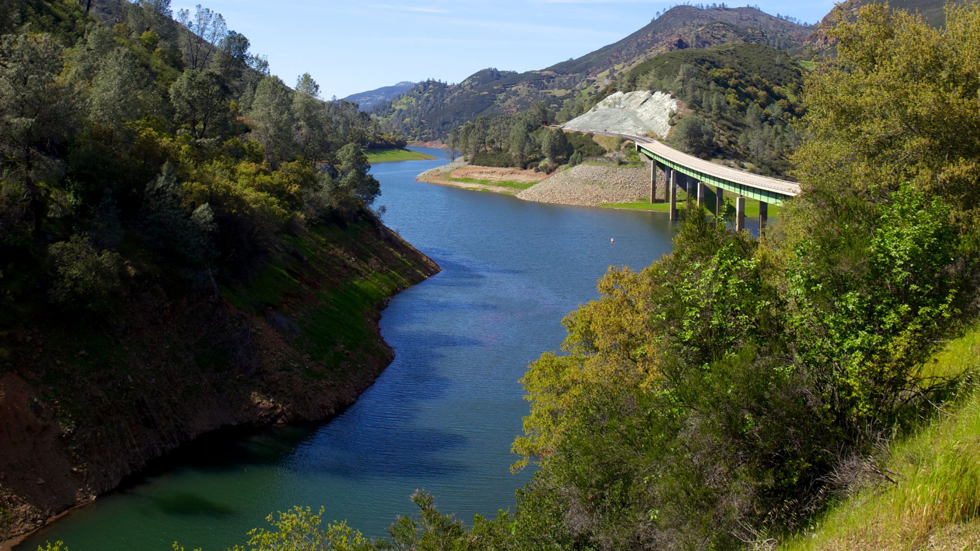 A scenic view of a large, blue-green lake winding through a valley, flanked by tree-covered hills. A multi-arched bridge, carrying California State Route 49, spans the lake in the distance, connecting the green hillsides. The sky above is clear and blue.