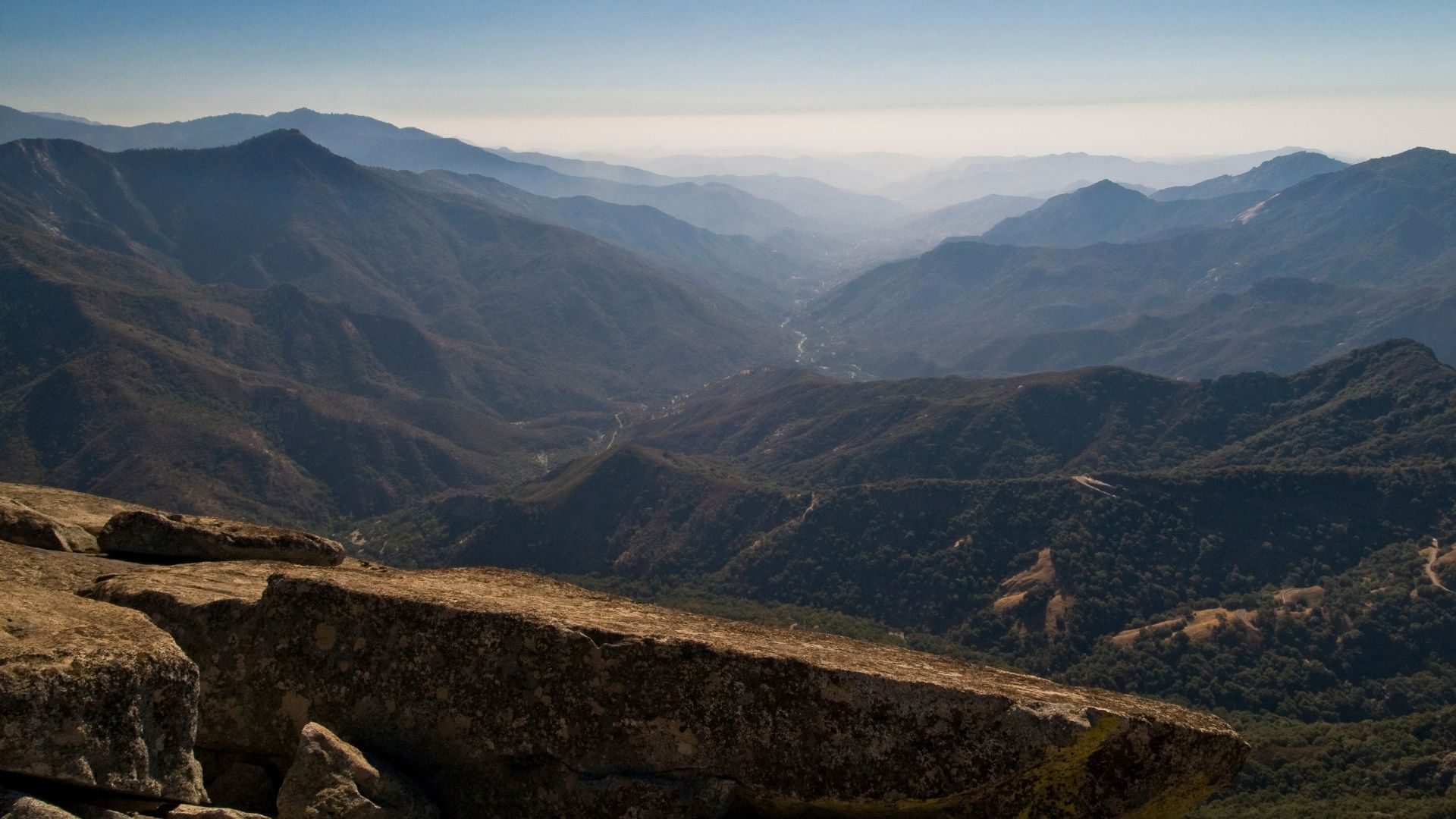A wide-angle view from a rocky outcrop overlooking a vast, hazy mountain valley with a winding river visible in the distance, all under a clear sky.
