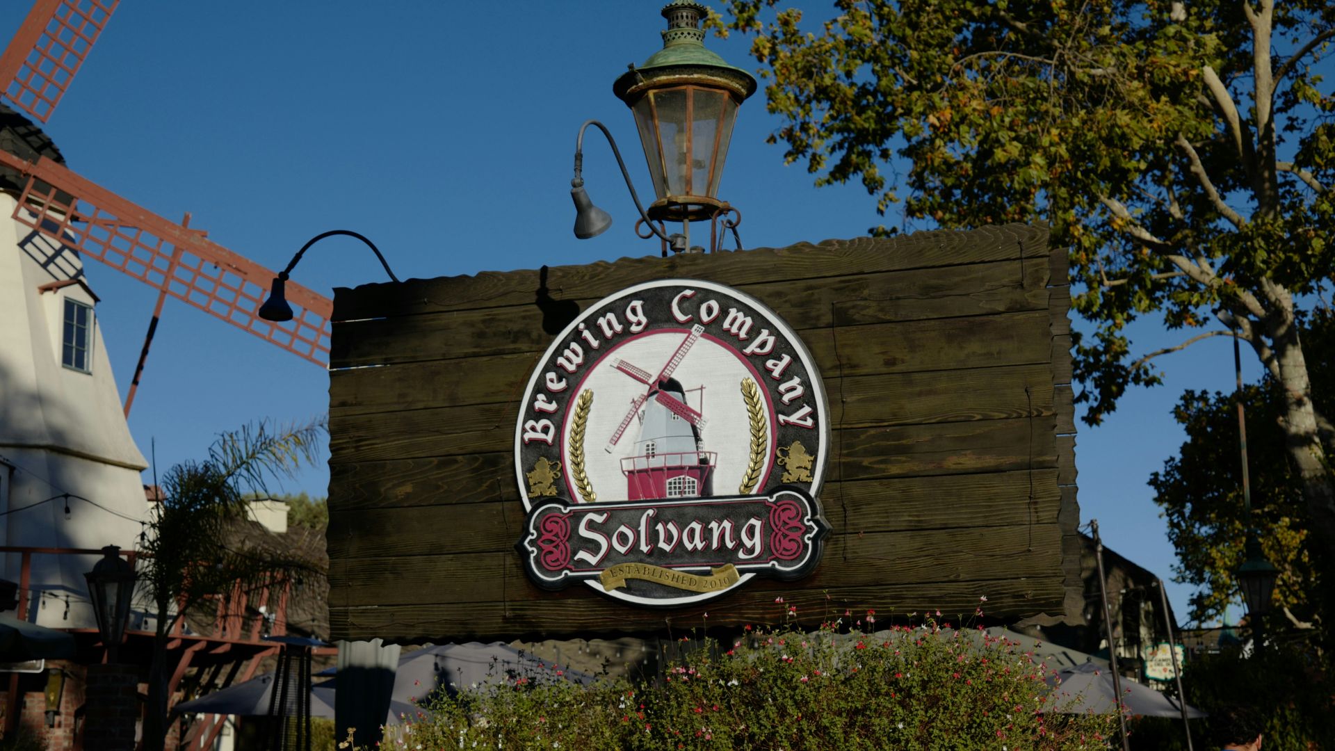 A wooden sign for Solvang Brewing Company featuring a windmill logo, set against a backdrop of buildings and greenery in the Danish-themed town of Solvang, California.