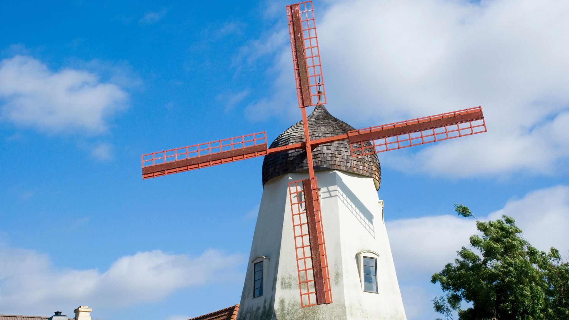 A white windmill with red blades and a brown shingled roof stands against a bright blue sky with scattered white clouds, in what appears to be the Danish-themed town of Solvang, California.