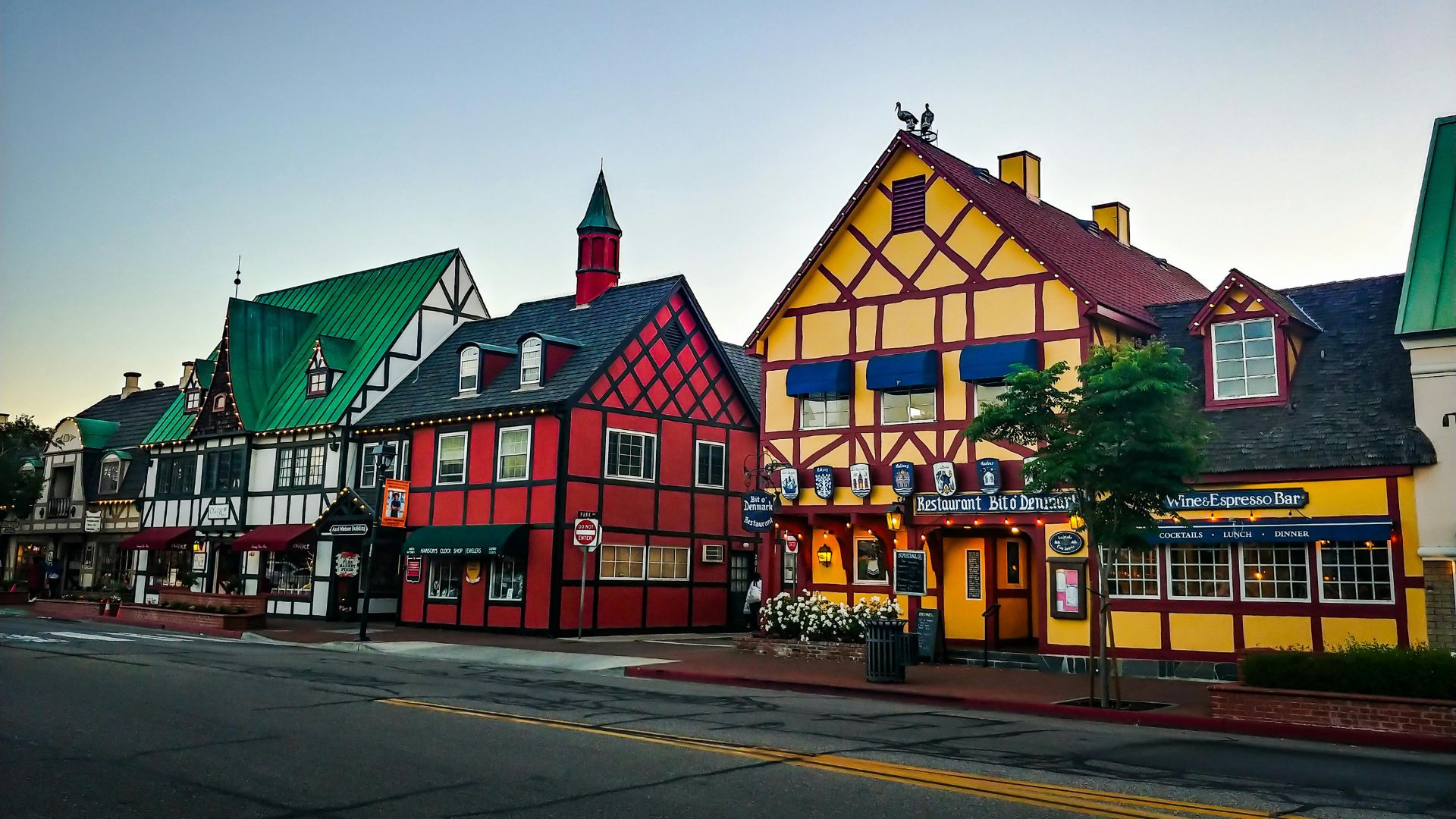 A street view of Solvang, California, featuring colorful buildings with Danish-style architecture, including half-timbered facades and steep gabled roofs, under a clear sky. The buildings house businesses like a restaurant and a wine & espresso bar, and the scene includes a street with markings and a tree.