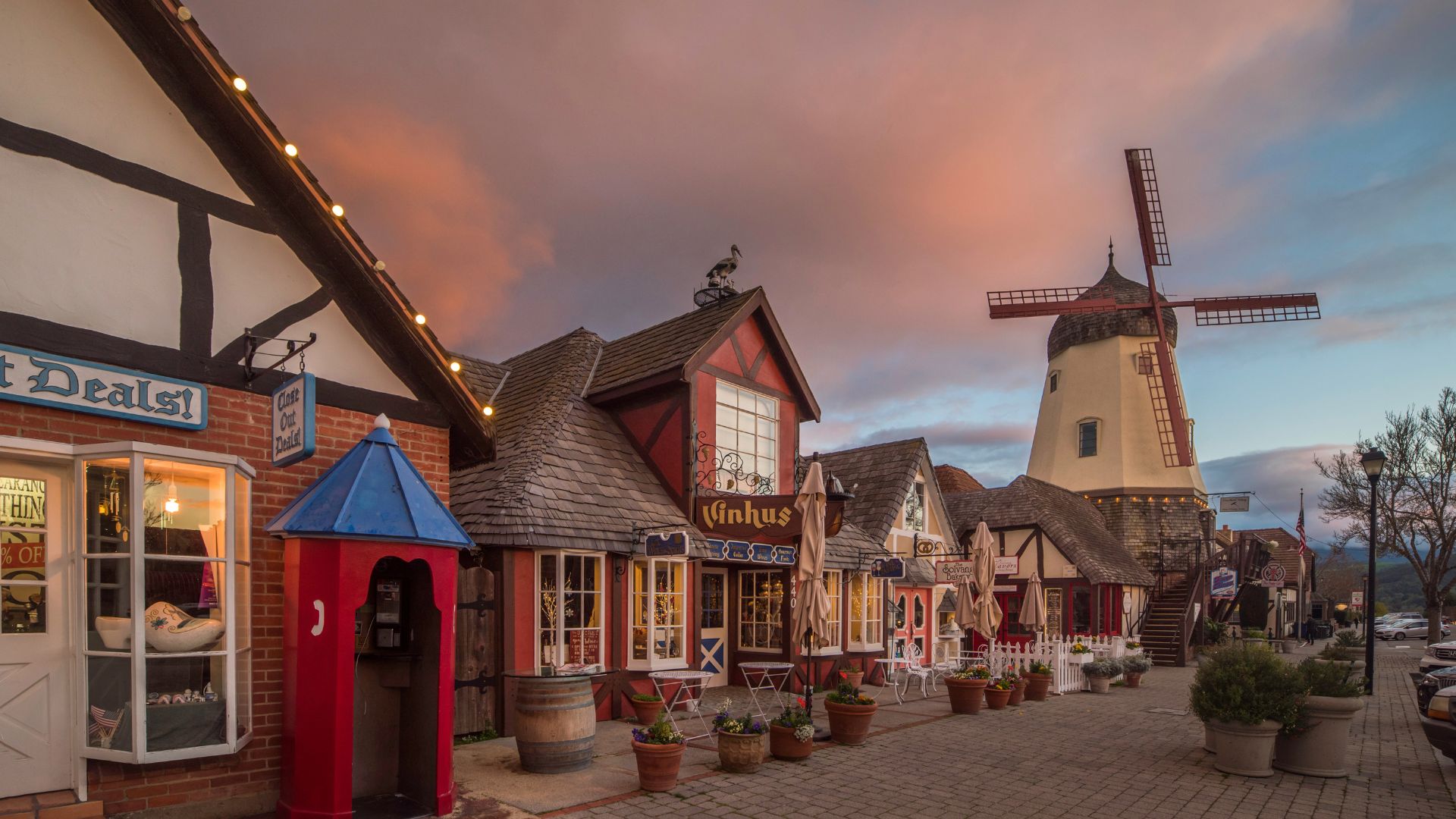 A nighttime view of a quaint street in Solvang, California, featuring buildings with Danish architectural elements, illuminated storefronts, a prominent windmill in the background, and crosswalks in the foreground, all under a cloudy sky. 